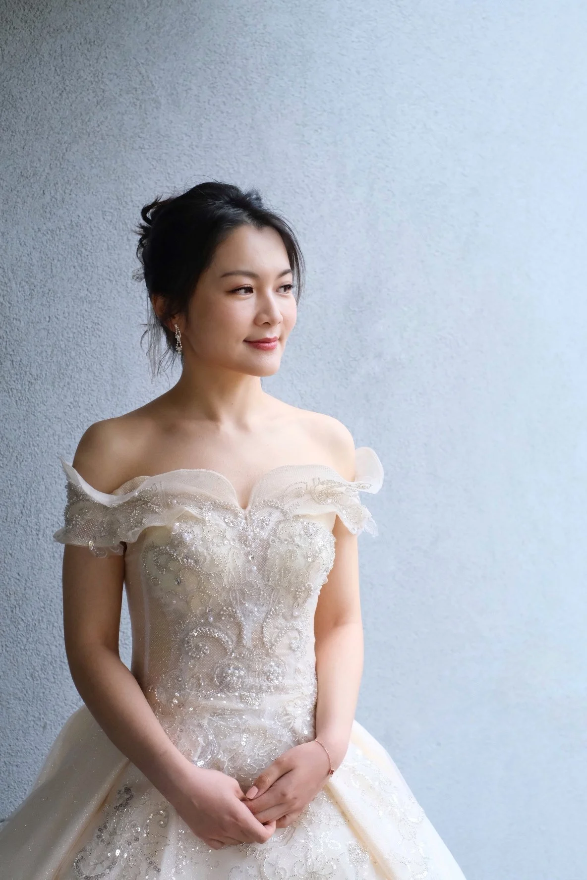 A woman wearing an elegant off-the-shoulder ivory wedding gown with intricate embroidery, standing against a textured gray wall, with her hands clasped in front of her.