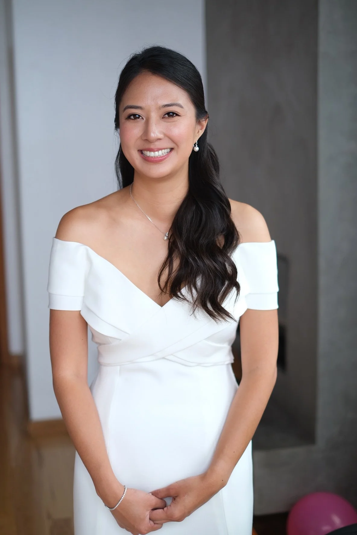 A smiling woman in a white off-the-shoulder dress standing indoors against a modern background.