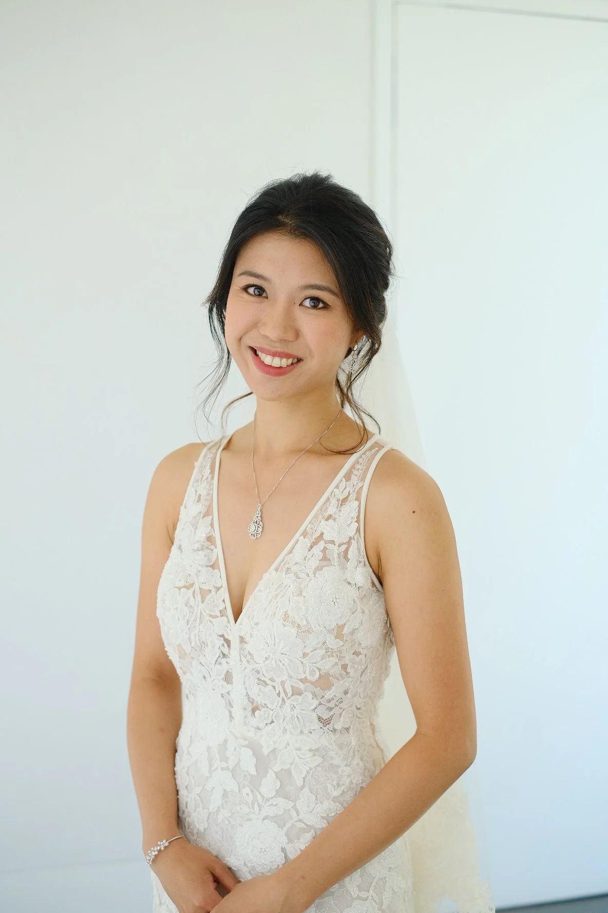 A woman in a white lace dress with floral patterns, wearing a silver necklace with a pendant and smiling with her hands clasped in front of her.