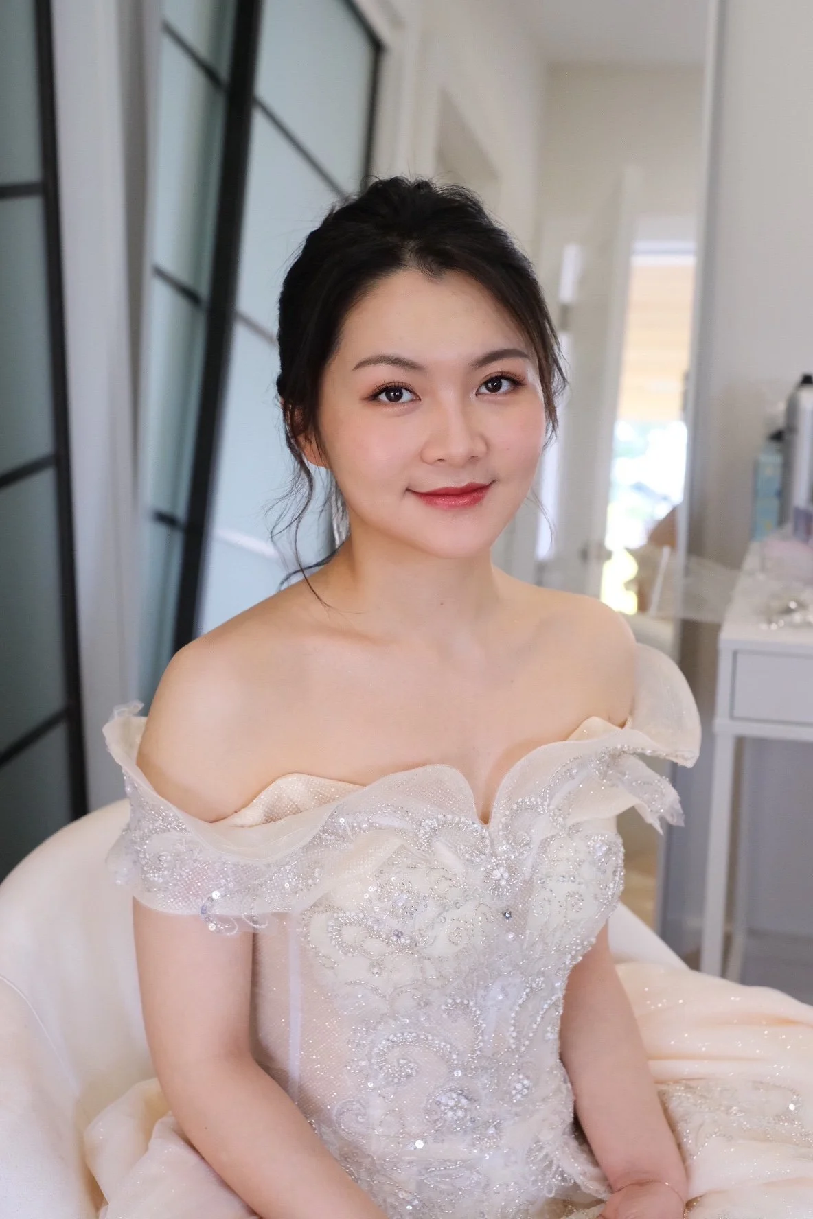 A young woman in an elegant white dress with intricate beadwork and ruffles, sitting indoors with a faint smile.