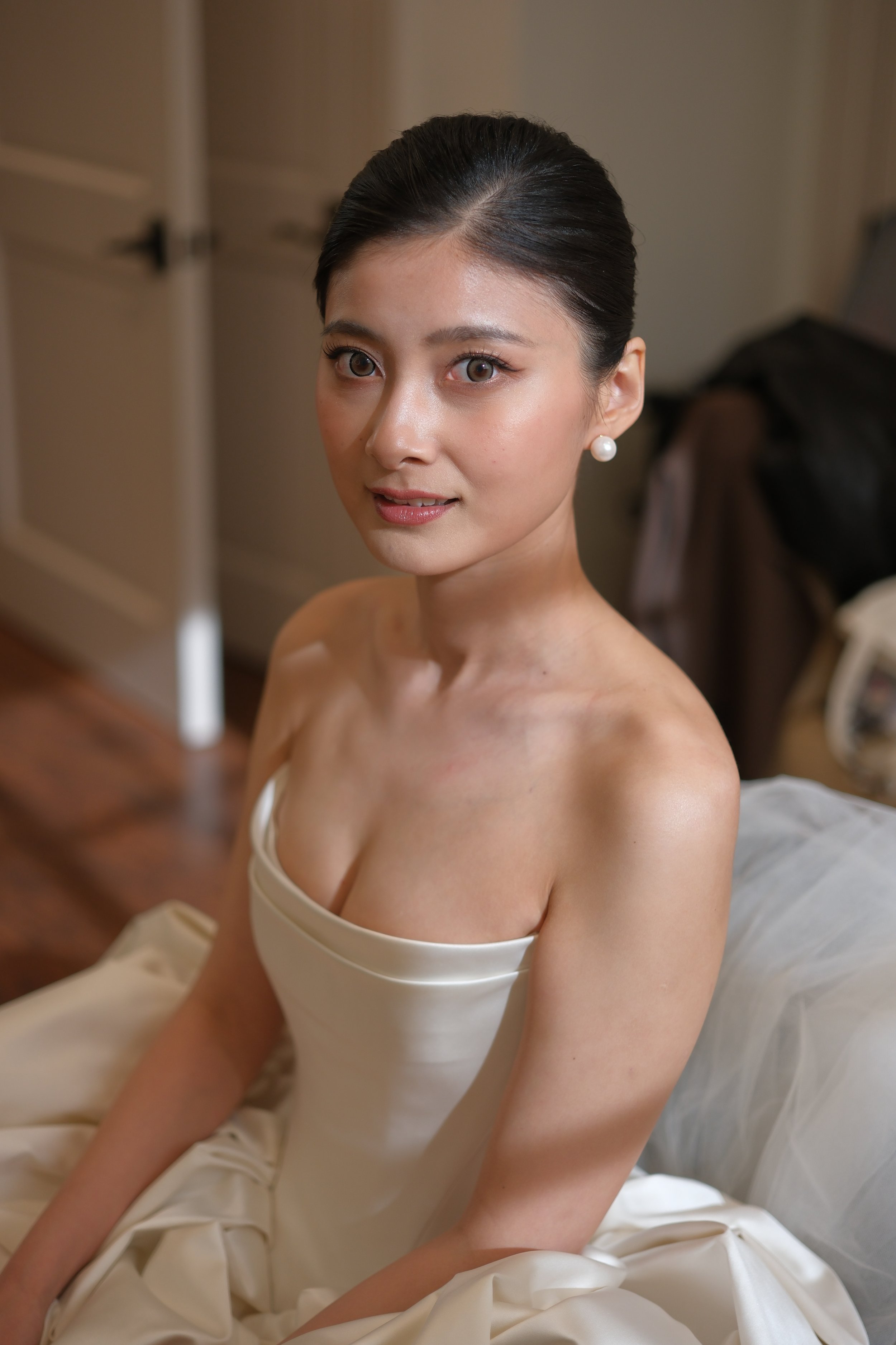 A woman with dark hair styled in an elegant updo, wearing pearl earrings and a strapless white dress, sitting on a bed with beige and white bedding in a softly lit room.