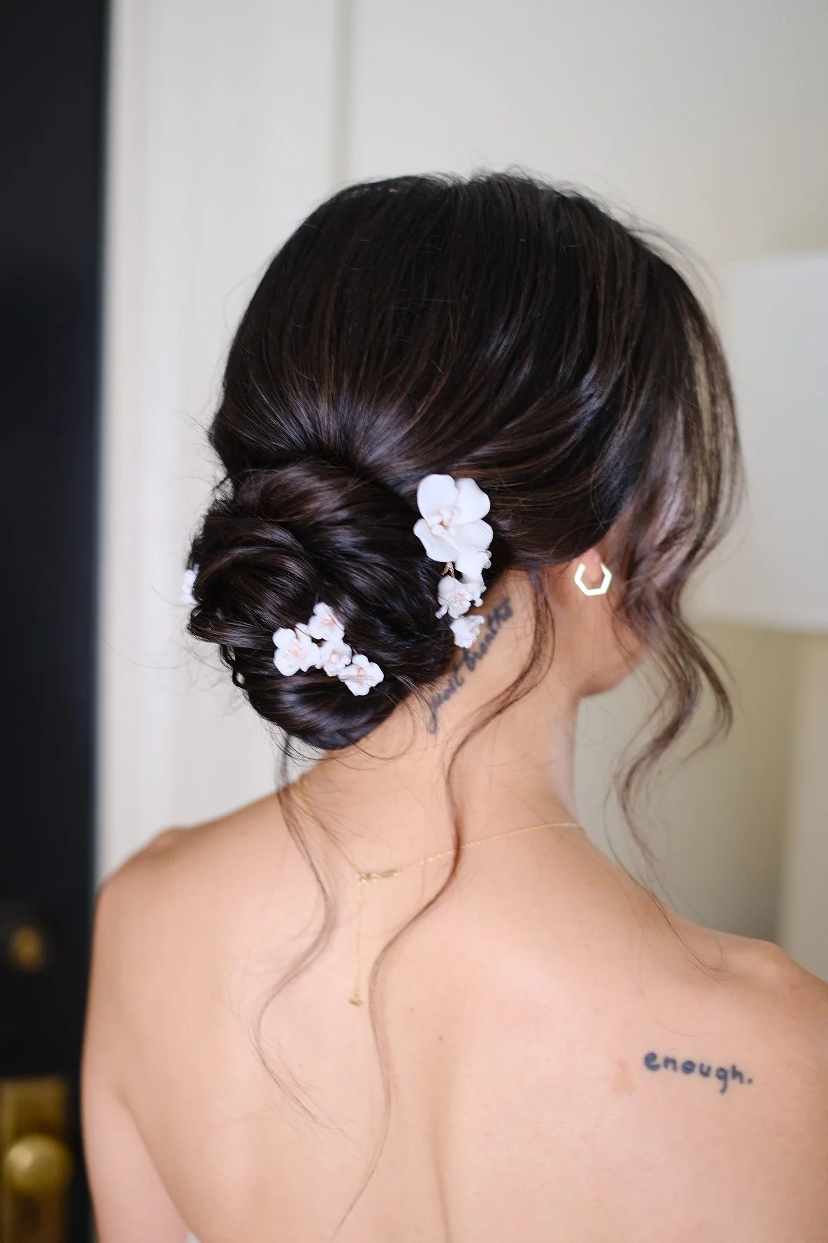 Back view of a woman with dark brown hair styled in an elegant updo adorned with white flowers, wearing gold hoop earrings and a delicate gold necklace, showing a tattoo reading 'enough.' on her shoulder.
