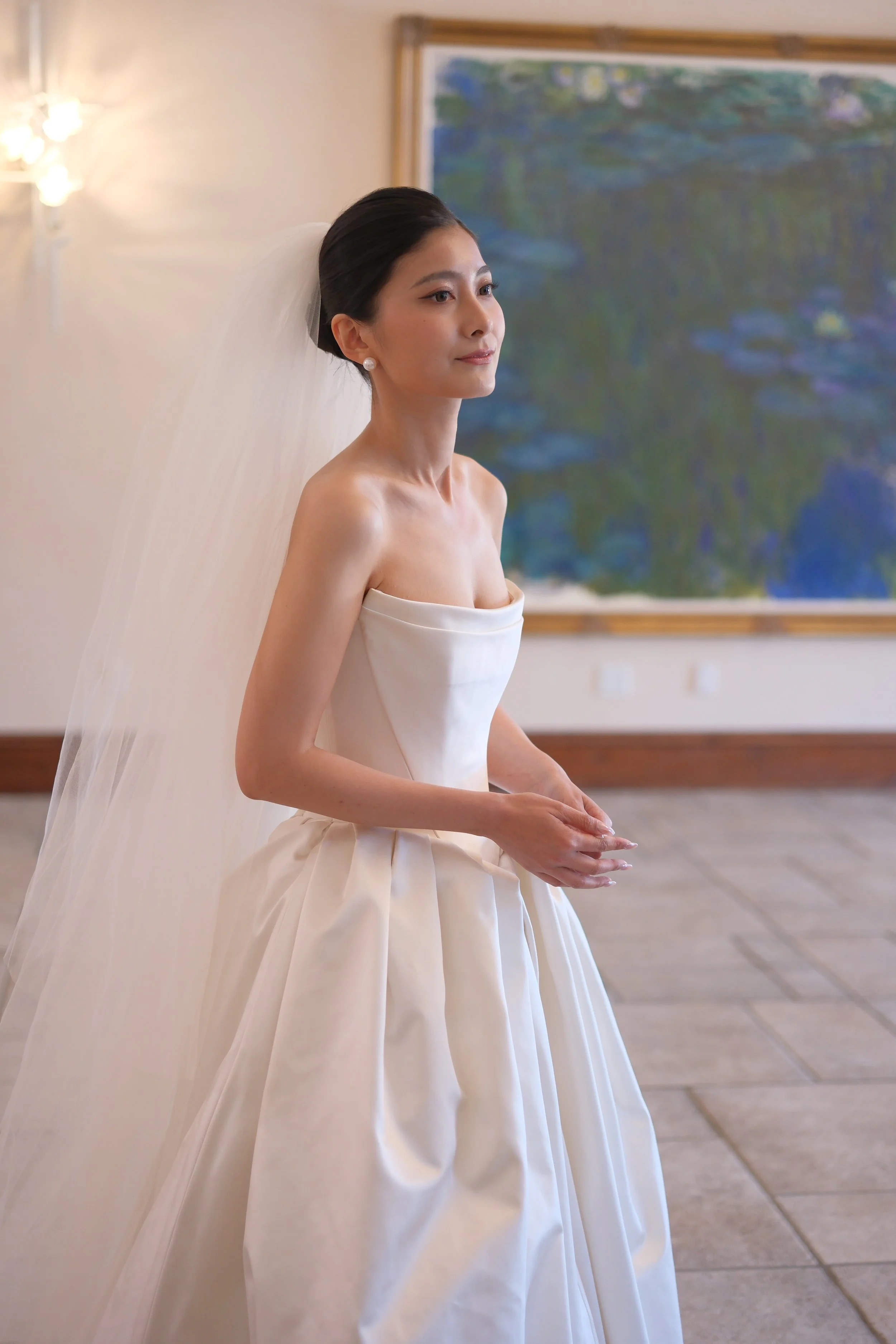 A woman in a white wedding dress with a veil, standing indoors with a large framed painting of water lilies in the background.
