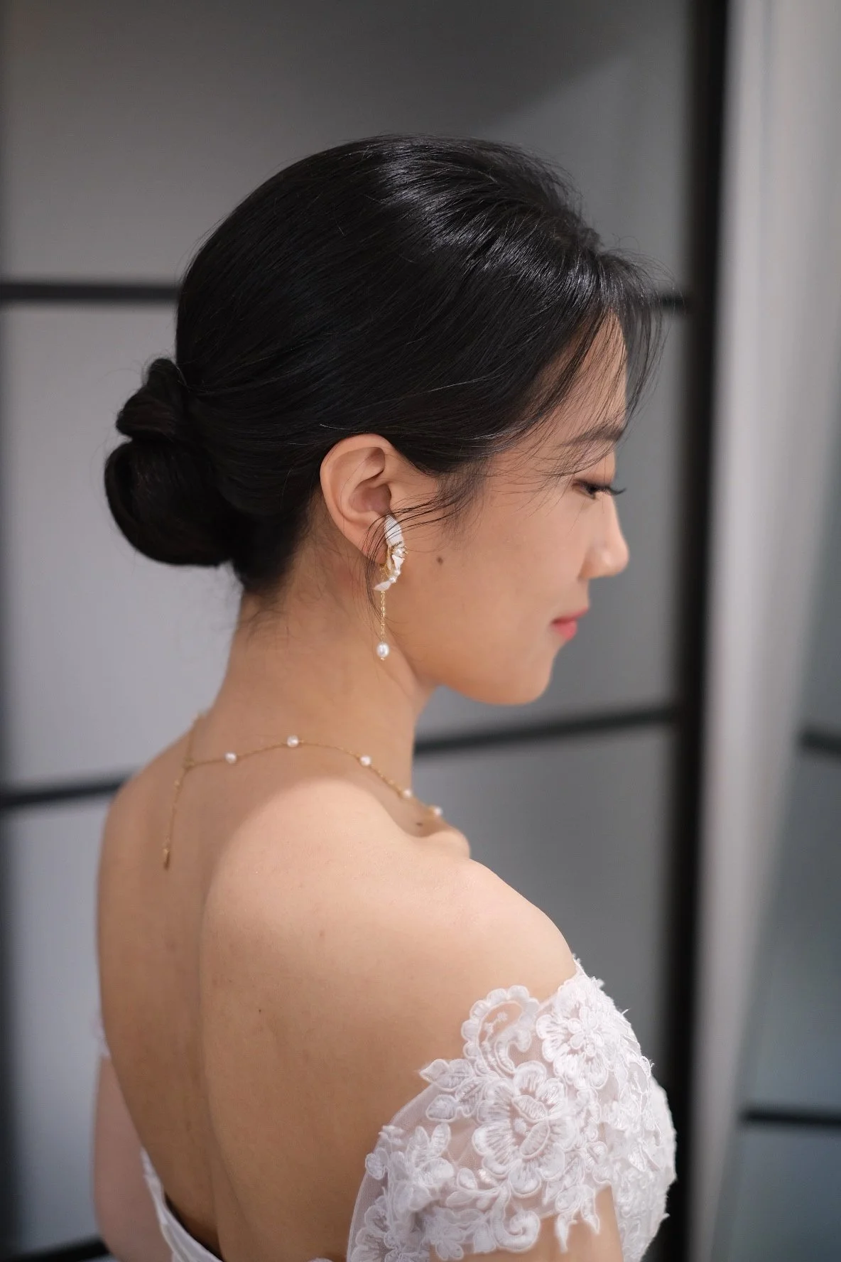 A woman with dark hair styled in a low bun, wearing elegant pearl and gold jewelry, posing in a white lace dress with off-the-shoulder sleeves.