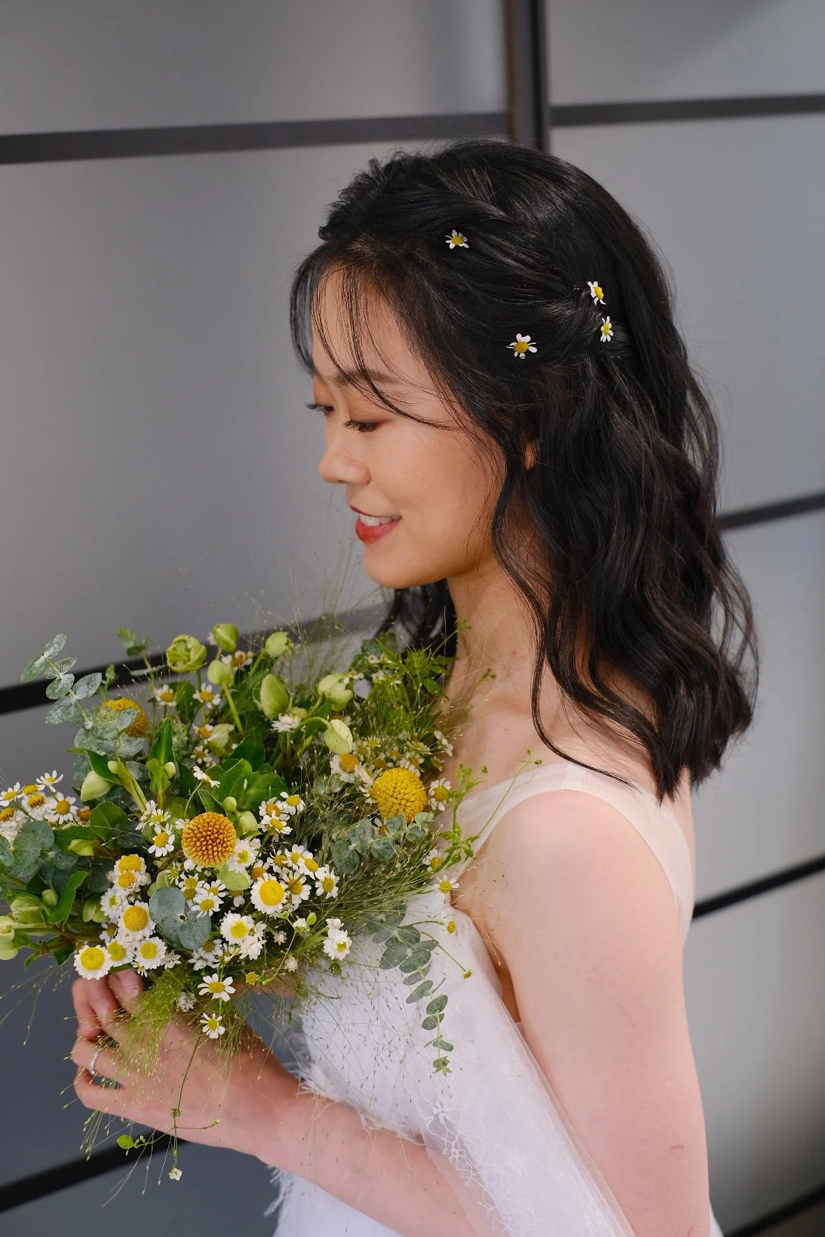 A woman in a white dress holding a bouquet of flowers, with small daisies in her dark, wavy hair, smiling gently.