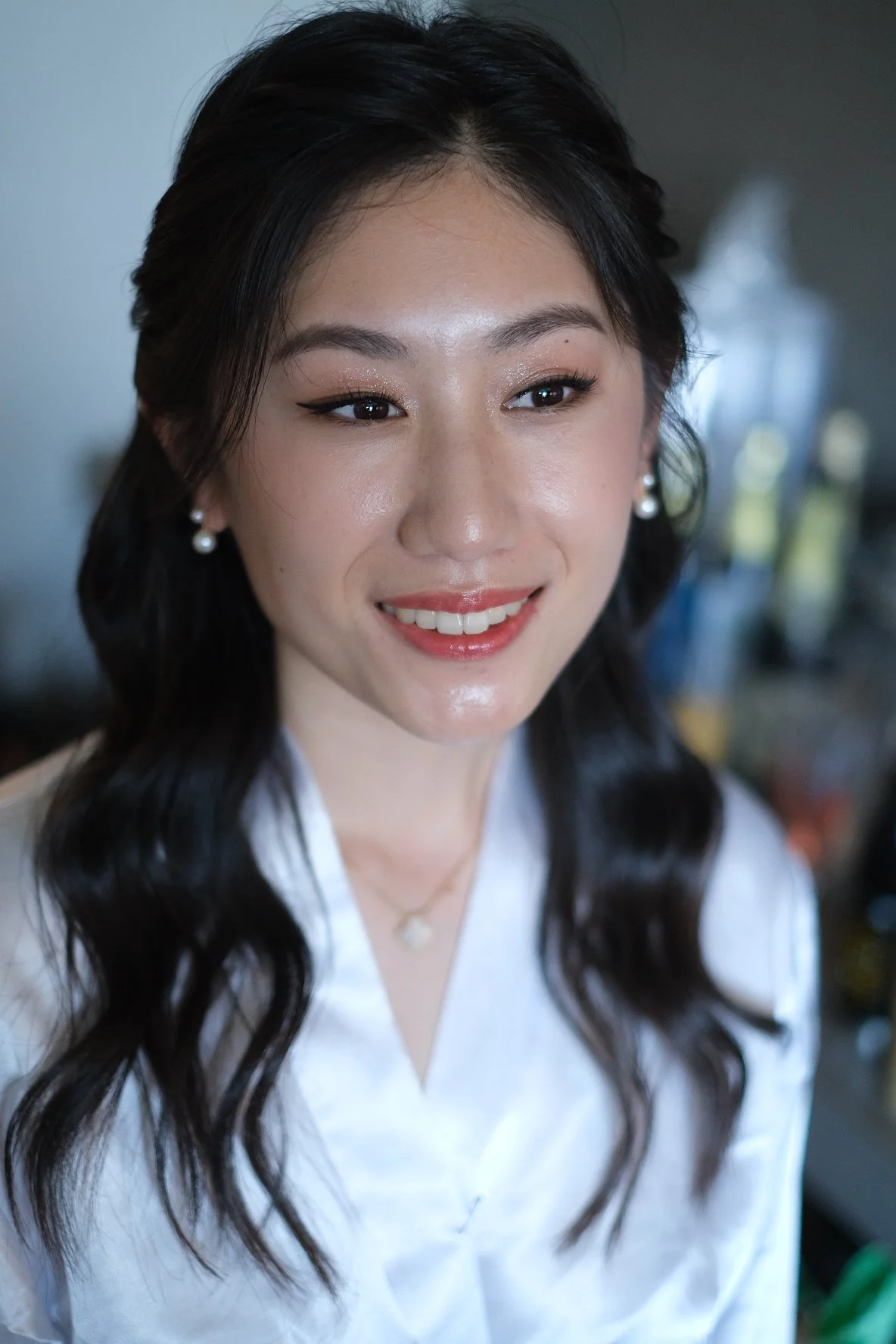 Close-up of a smiling woman with dark hair, pearl earrings, and light makeup, wearing a white top.