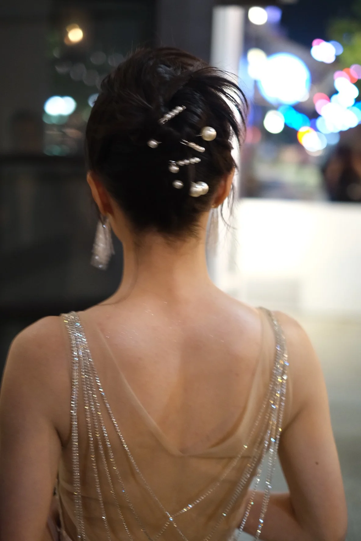 Back view of a woman with dark hair styled up with pearl and metallic hairpins, wearing a glamorous, sleeveless, beige dress with sparkly embellishments.