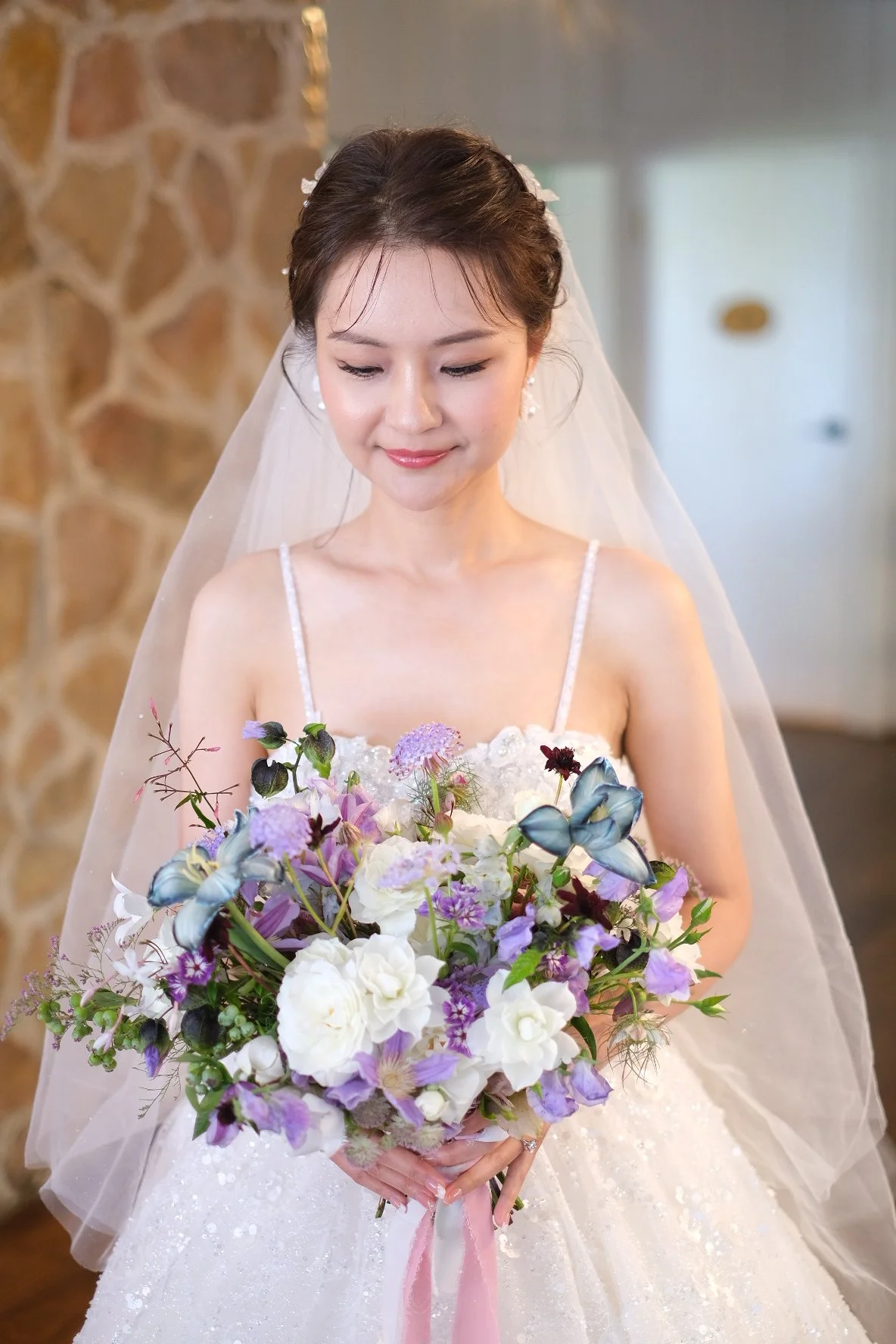 A bride in a wedding dress holding a bouquet of purple, white, and green flowers, with a sheer veil and smiling gently.