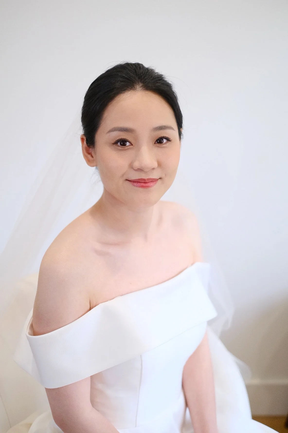 A portrait of a young woman with dark hair, wearing a white off-the-shoulder dress, sitting against a white background, smiling gently.