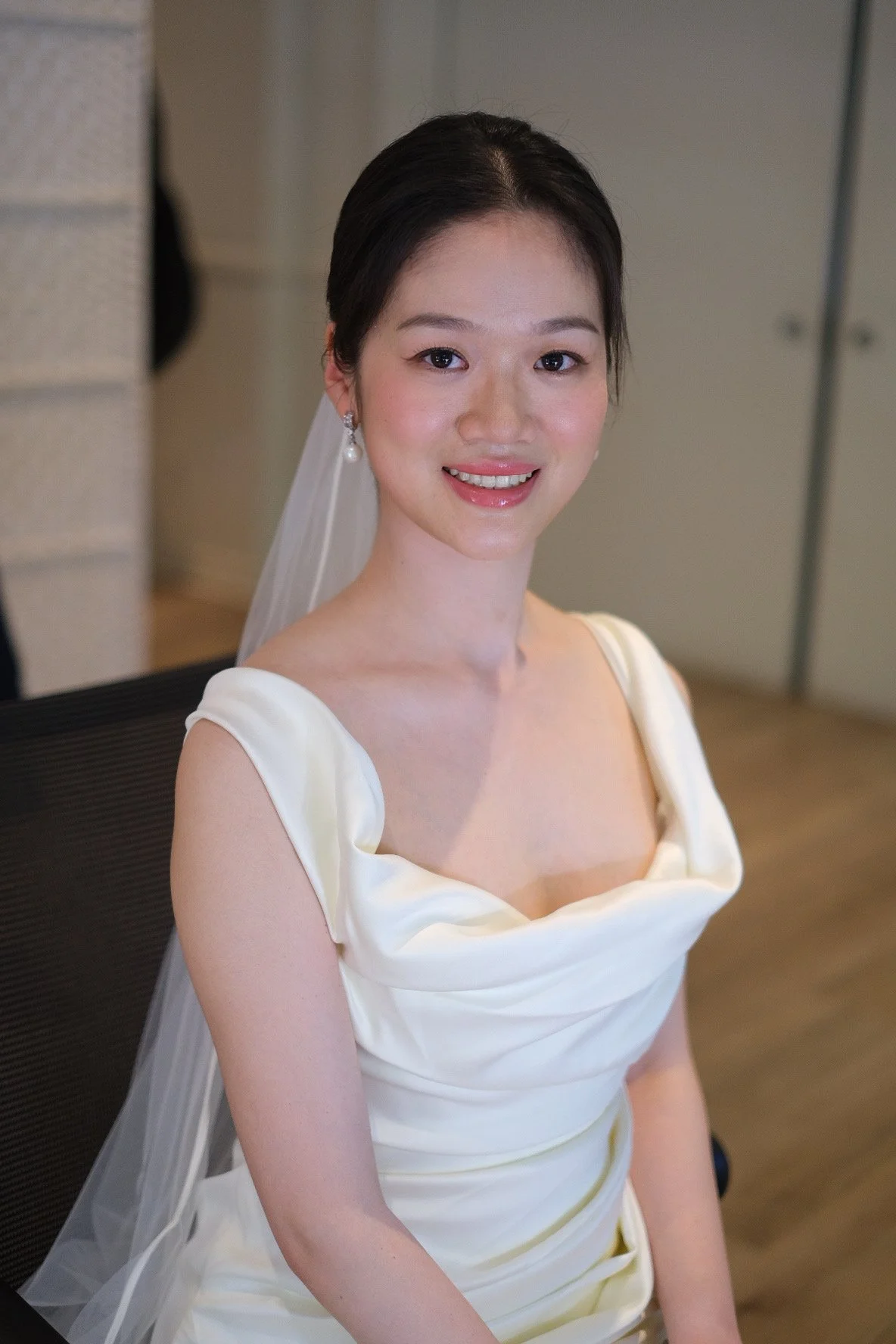 A smiling young woman with dark hair styled in an updo, wearing pearl earrings and a white satin wedding dress with a dramatic neckline, sitting on a black chair indoors.