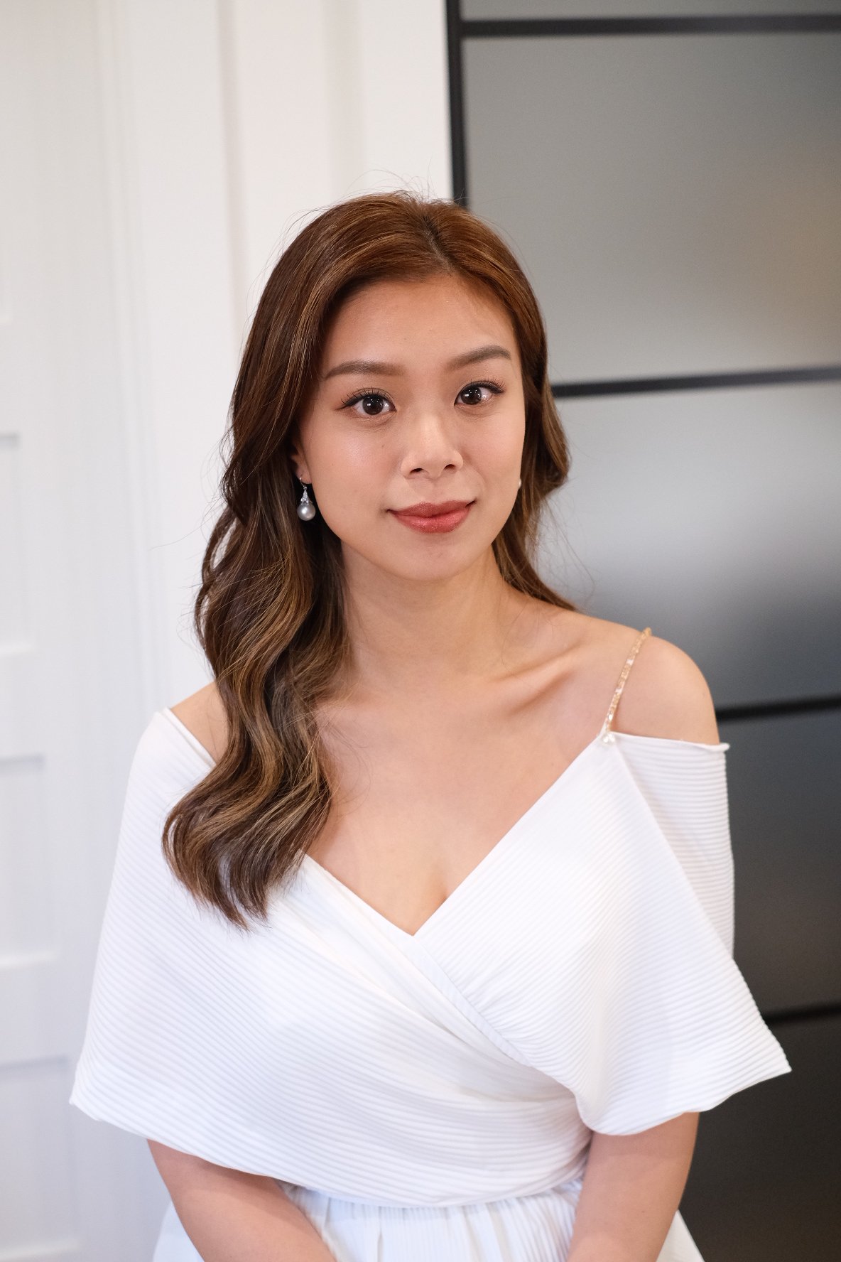 A portrait of a woman with long wavy brown hair wearing pearl earrings and a white off-shoulder dress, standing indoors with a neutral background.