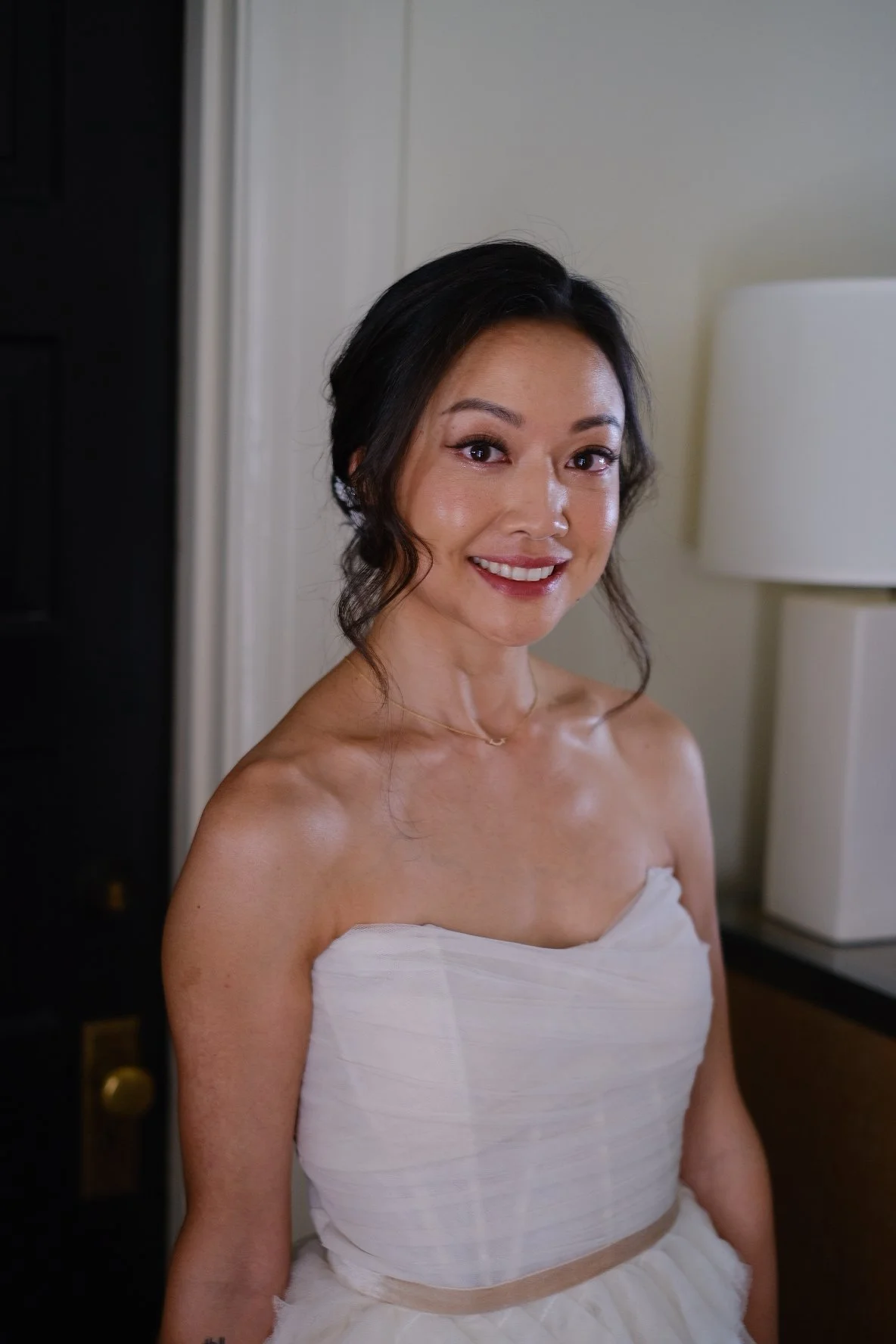 A woman with dark hair styled in loose waves smiling at the camera, wearing a strapless white dress with a ruched bodice and a fluffy skirt, standing indoors near a white wall and a white lamp.