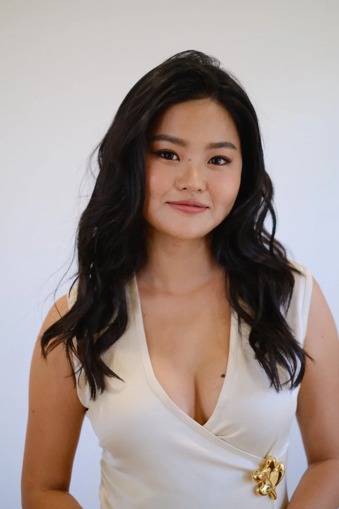 A young woman with long, dark, wavy hair and light skin, wearing a sleeveless cream-colored dress with decorative gold embellishment on the waist, standing against a plain white wall.