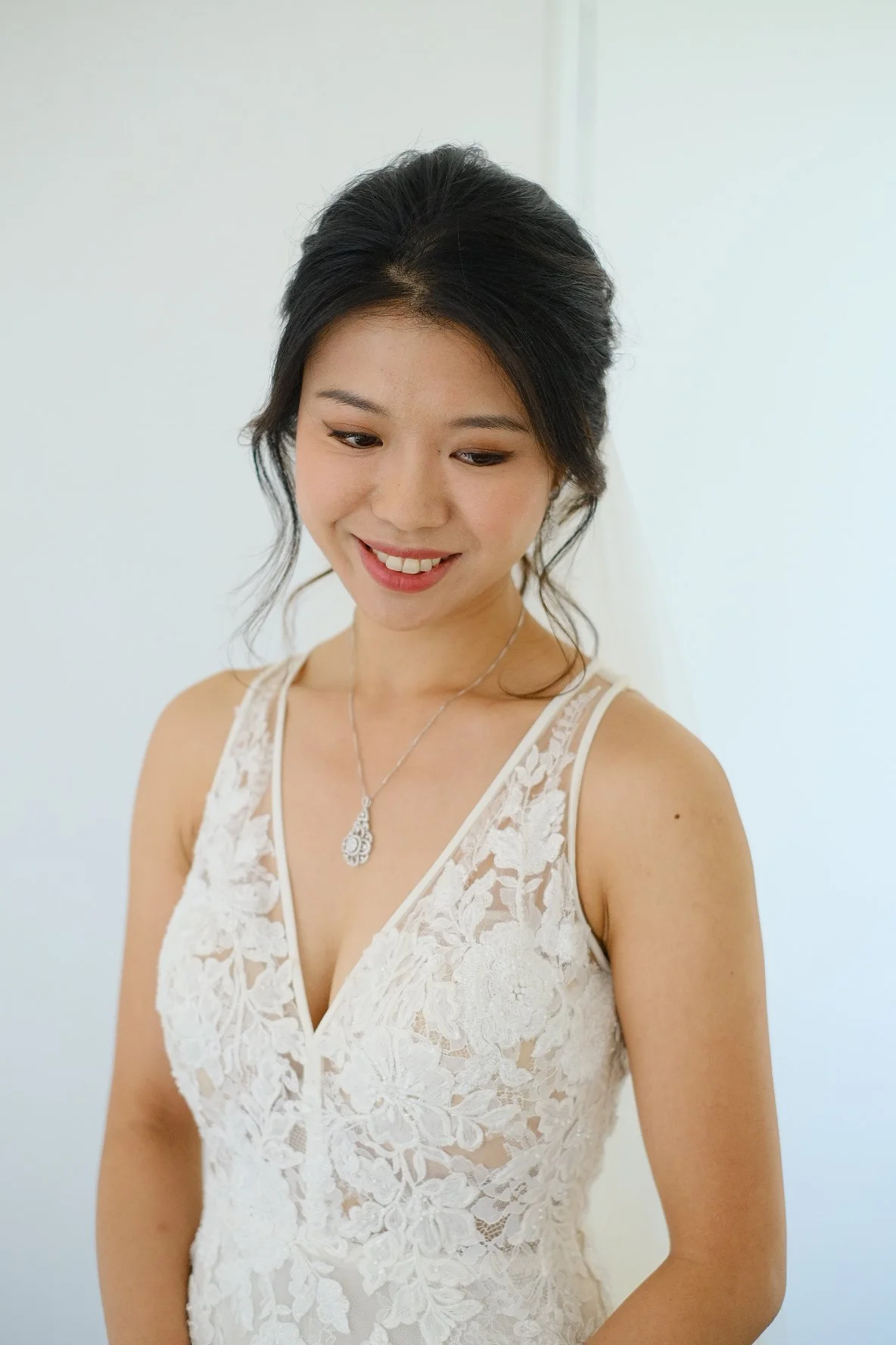 A young woman in a white lace dress with a necklace, smiling and looking downward.