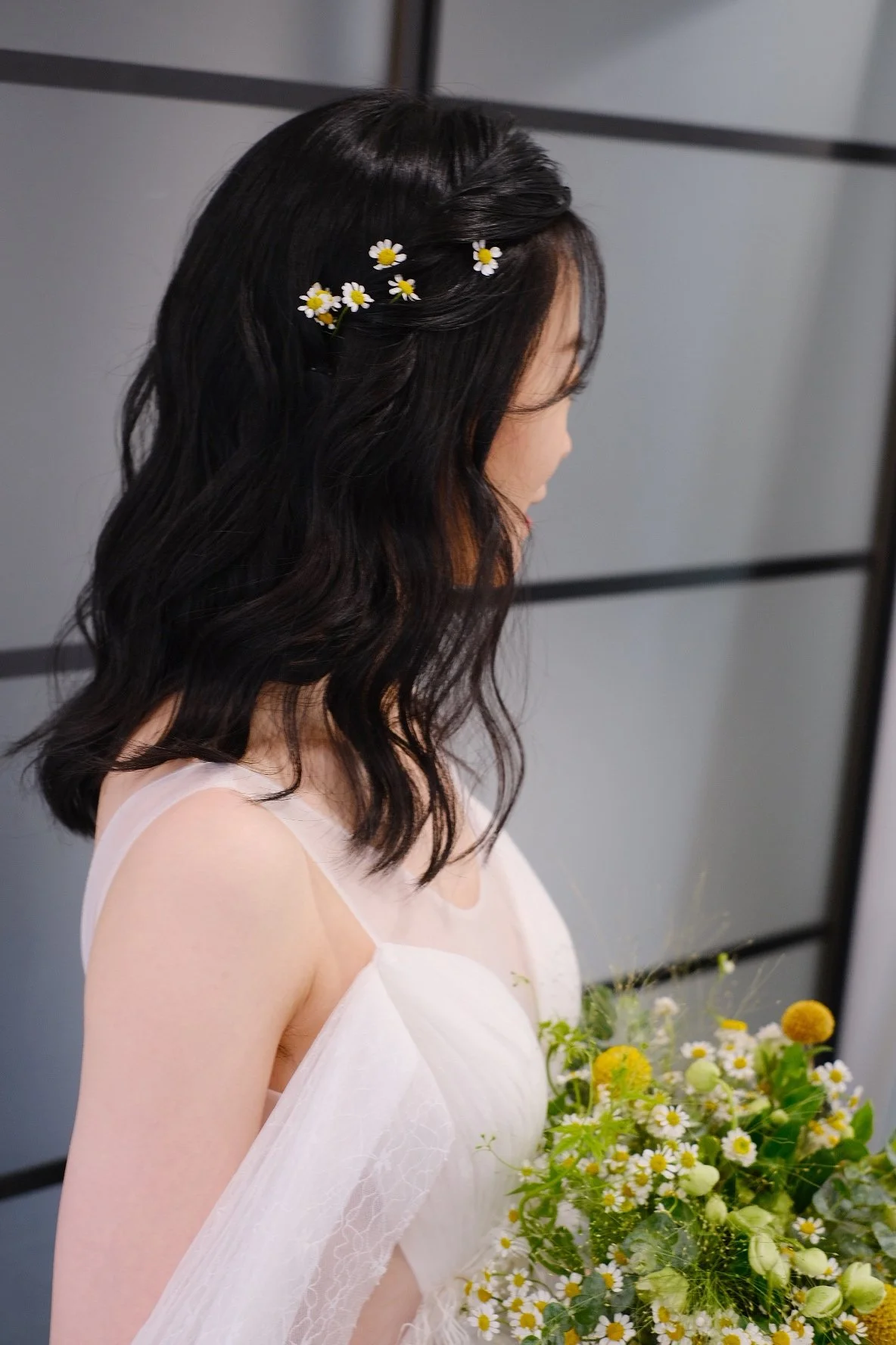 A woman with dark wavy hair decorated with small white daisies, wearing a white dress, holding a bouquet of white and yellow flowers, standing against a gray background.