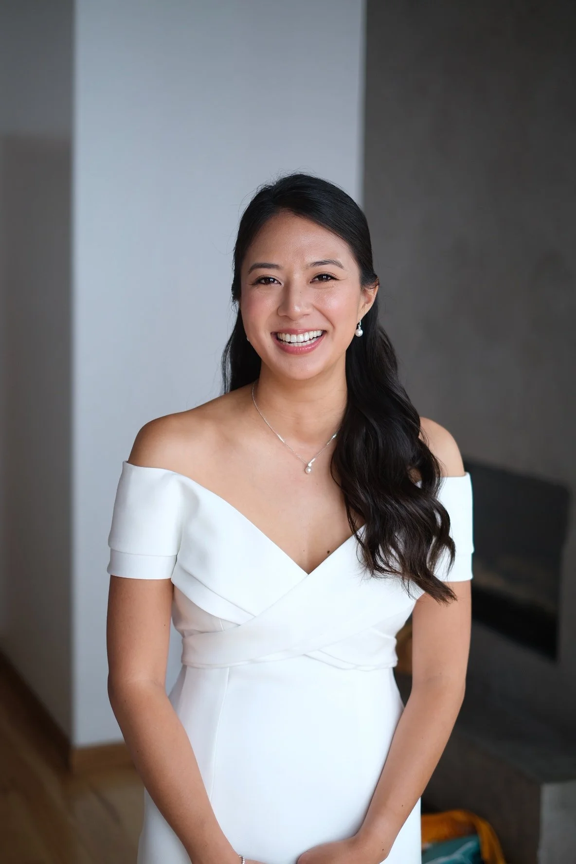 A smiling woman in an off-the-shoulder white dress standing indoors against a gray and white background.