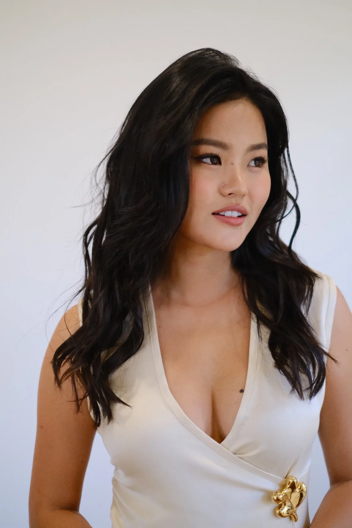 Portrait of a young woman with long black wavy hair, wearing a cream-colored sleeveless top with a small gold decorative detail on the left side, standing against a plain white background.