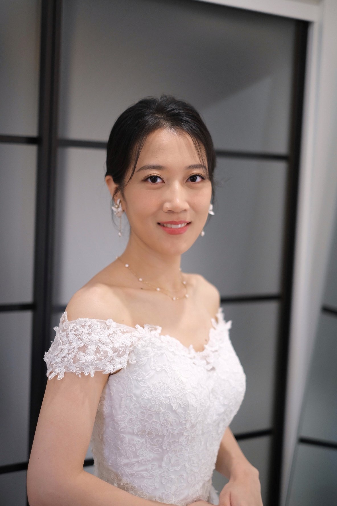 Woman wearing a white lace wedding dress with off-the-shoulder sleeves, standing indoors near a black partition, smiling at the camera.