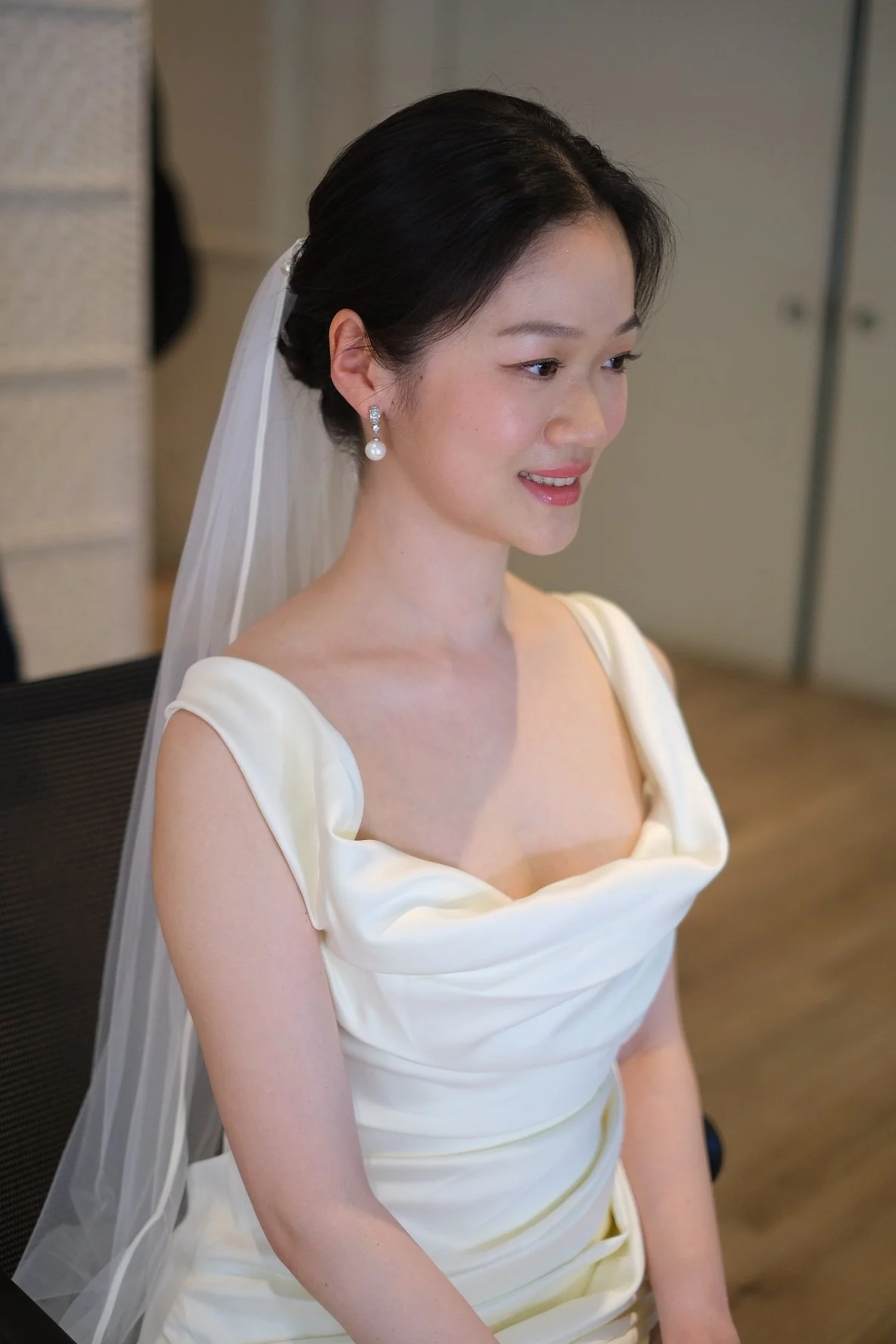 A woman in a white wedding dress with a veil and pearl earrings, sitting indoors with a slight smile.