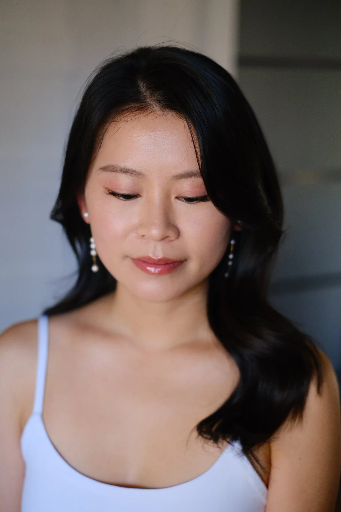 A woman with long black hair wearing a white sleeveless top and pearl earrings, looking down with a gentle smile.