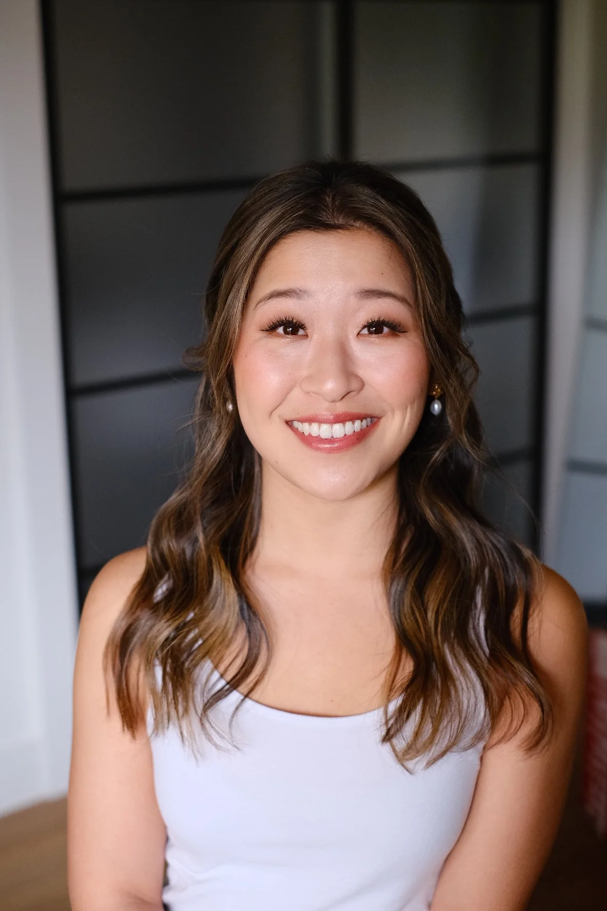 A young woman with long wavy brown hair, wearing pearl earrings and a white sleeveless top, smiling in a well-lit indoor setting with black shelving in the background.