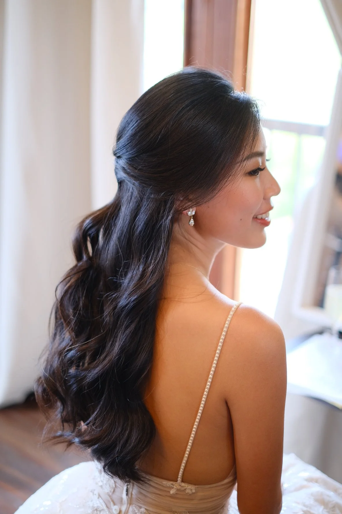 A woman with long, wavy dark hair styled in loose curls, wearing pearl earrings and a spaghetti strap dress, sitting near a window with curtains, smiling gently.