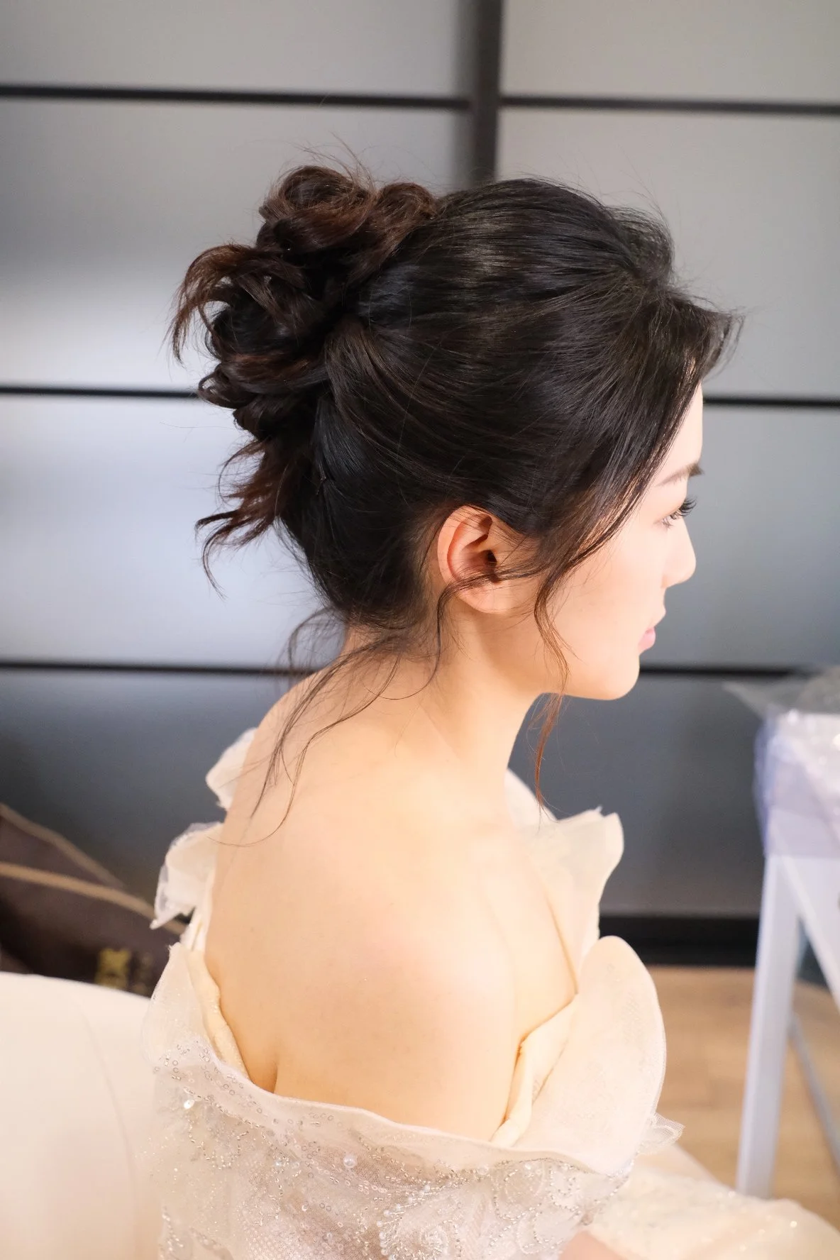 Side view of a woman with dark, wavy hair styled in an elegant updo, dressed in a light-colored, strapless dress with lace details.