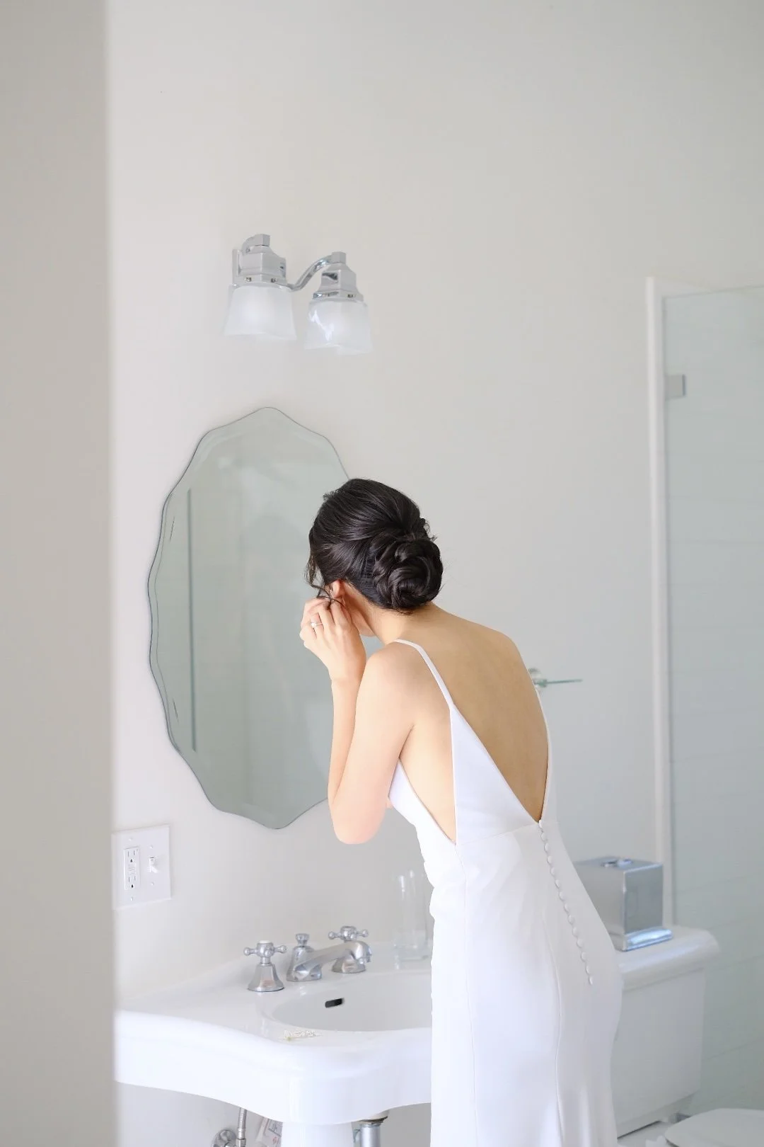 A woman in a white dress adjusting her earring in front of a mirror in a bathroom, with a white sink and a wall-mounted light fixture above the mirror.