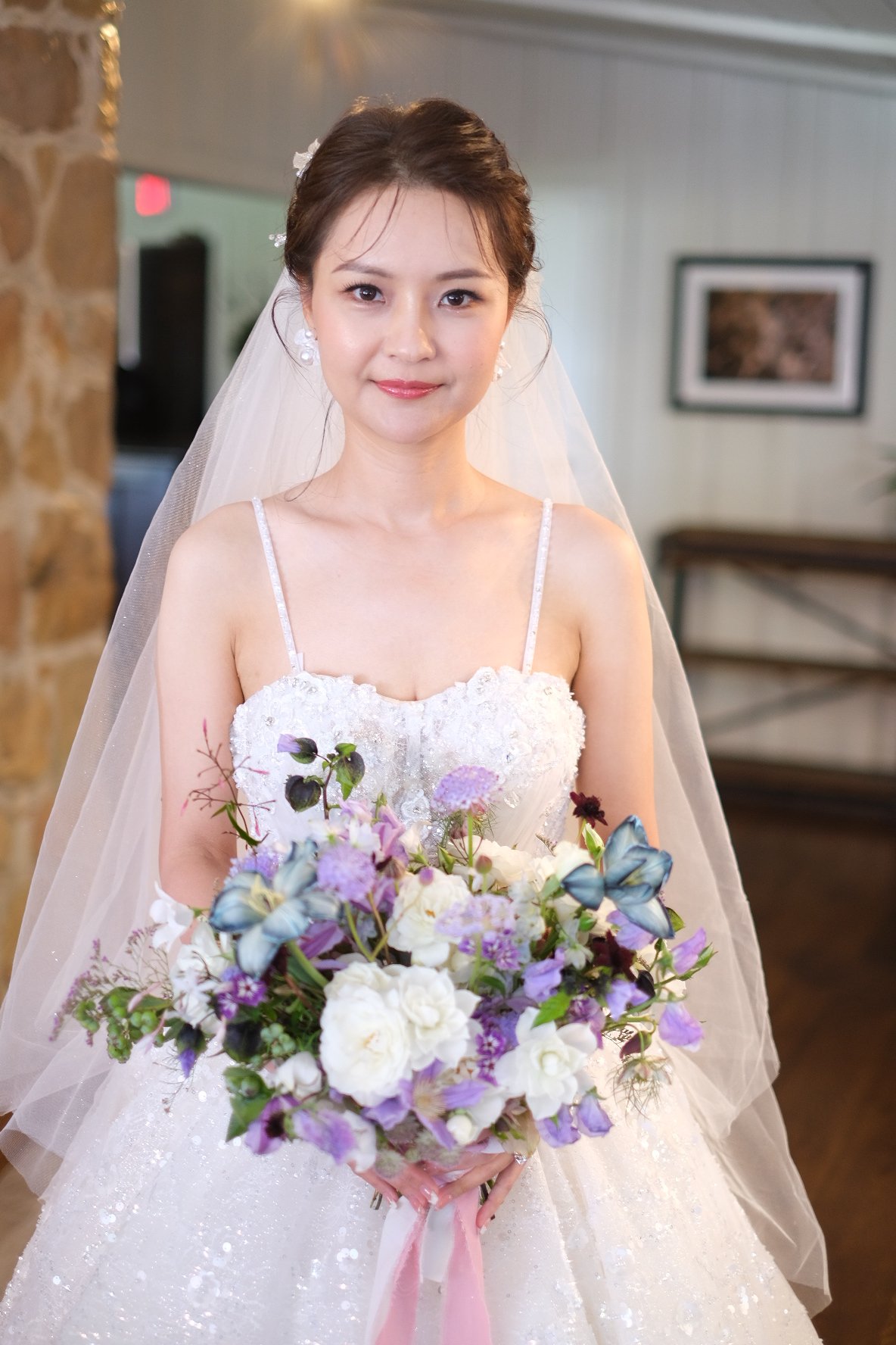 A bride in a white wedding dress holding a bouquet of purple, white, and green flowers with blue butterflies, standing indoors in front of a stone wall and framed pictures.