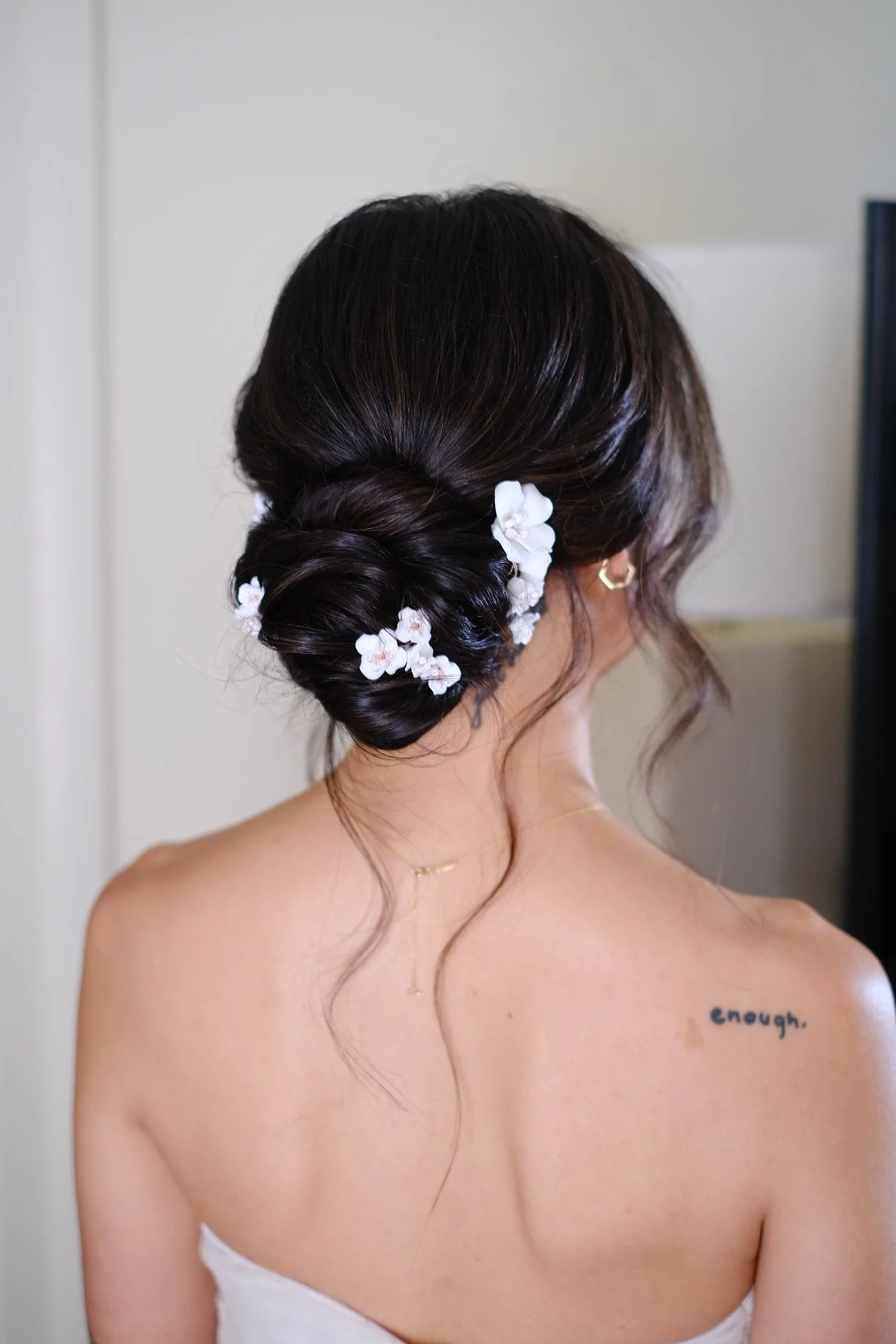 Back view of a woman with dark hair styled in an elegant updo, decorated with white flowers, wearing gold jewelry, and a white strapless dress, showing a tattoo on her upper back that reads 'enough'.