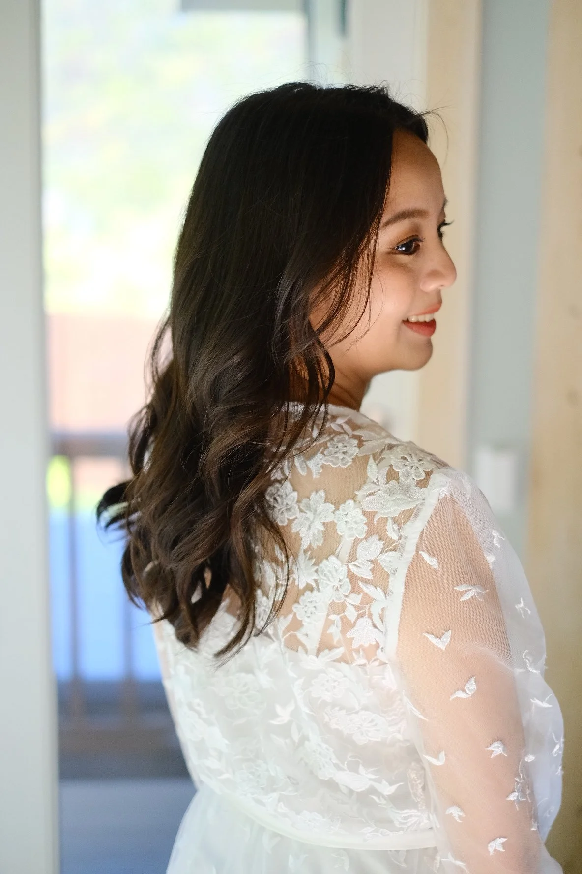 A woman with long, dark, wavy hair wearing a white lace dress with floral embroidery, smiling while standing indoors near a door with light coming through a window behind her.