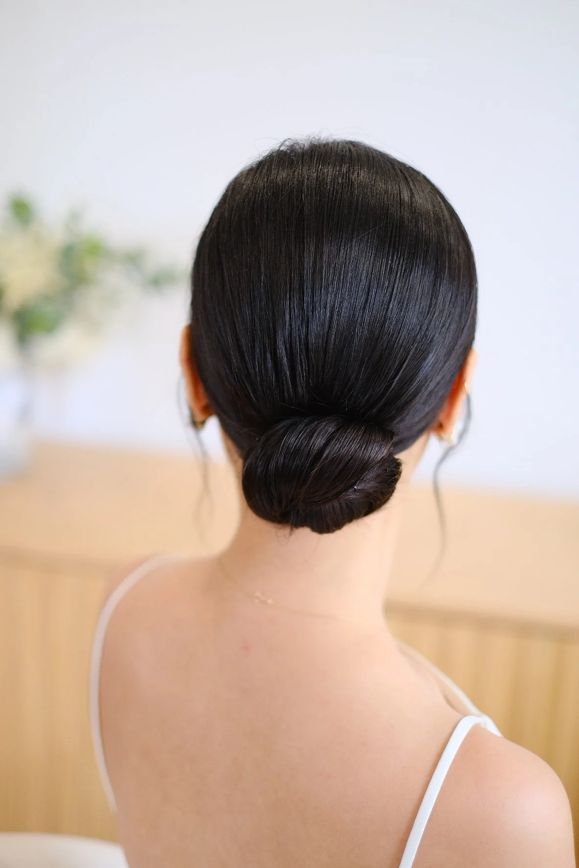 The back of a woman's head with sleek, shiny black hair styled into a low bun. She wears a white spaghetti strap top, with a blurred background that includes some green plants.
