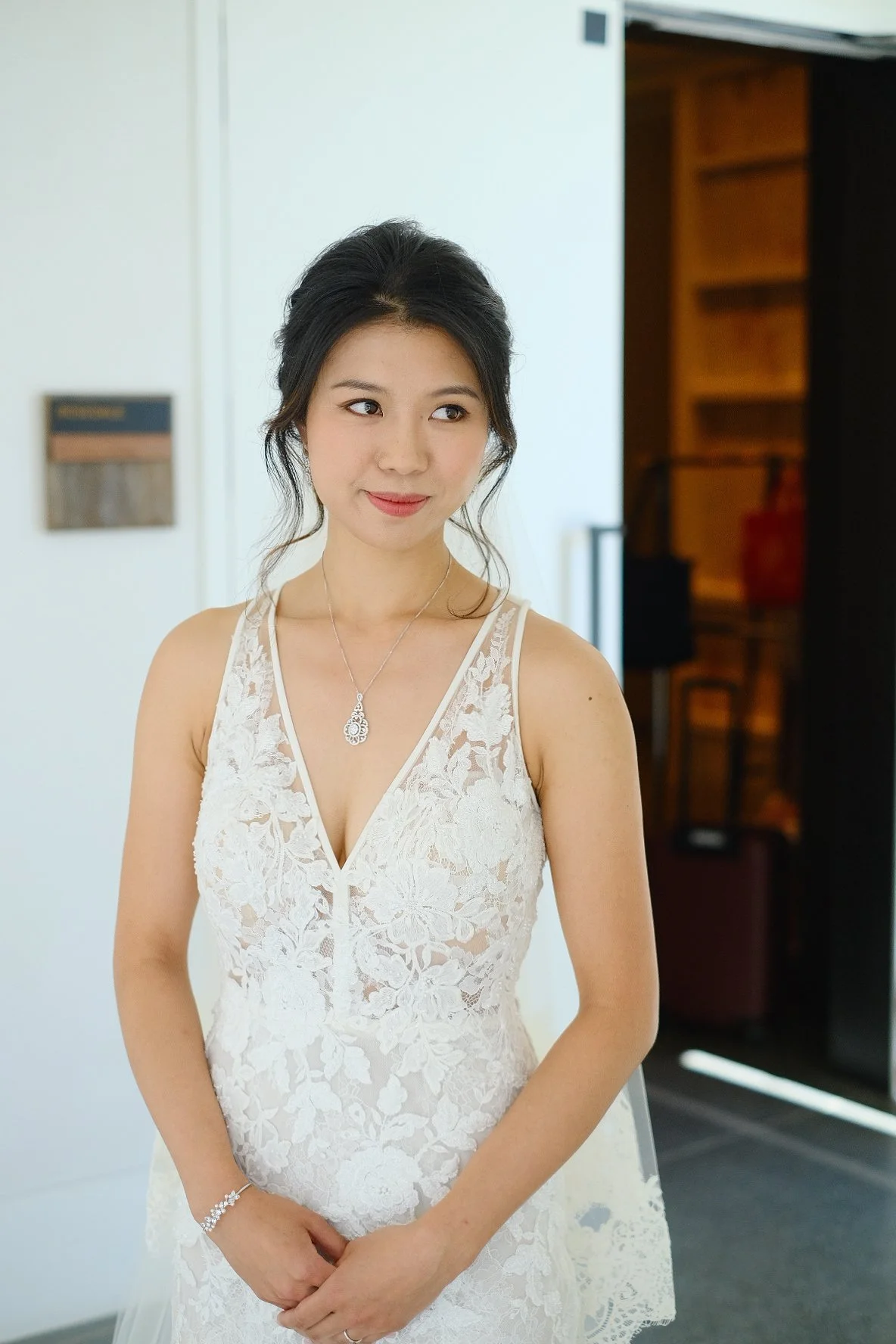 A woman in a white lace dress with a V-neckline and a pendant necklace, standing indoors with a neutral expression.