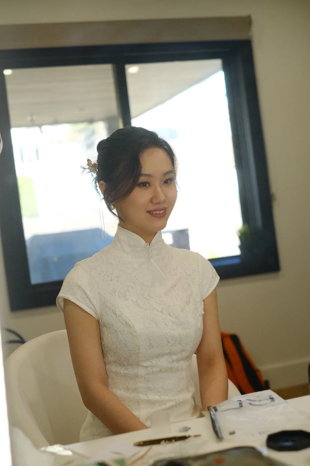 A young woman with dark hair styled in an updo, wearing a white lace dress with a high collar, sitting in a room with a large window in the background.