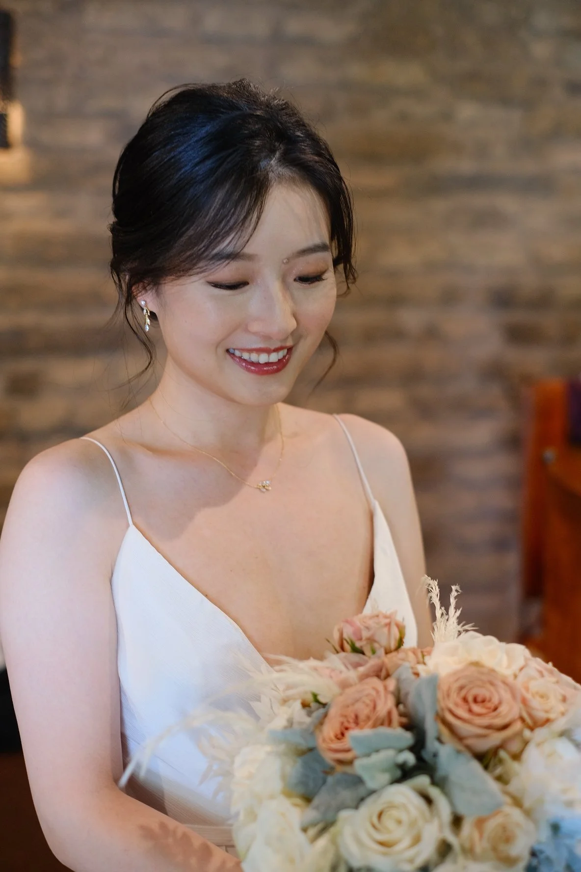 A smiling woman in a white dress holding a bouquet of pink, white, and cream roses, with a blurred brick wall background.