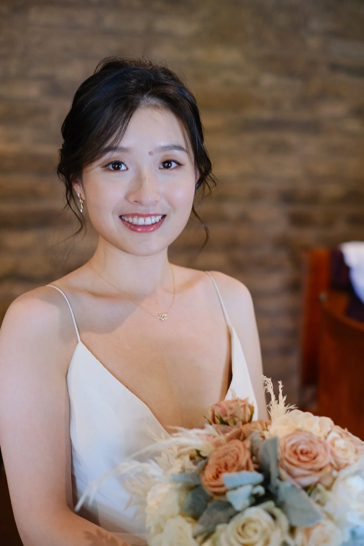 A woman in a white dress is smiling and holding a bouquet of pink and white flowers, standing indoors with a wooden wall background.