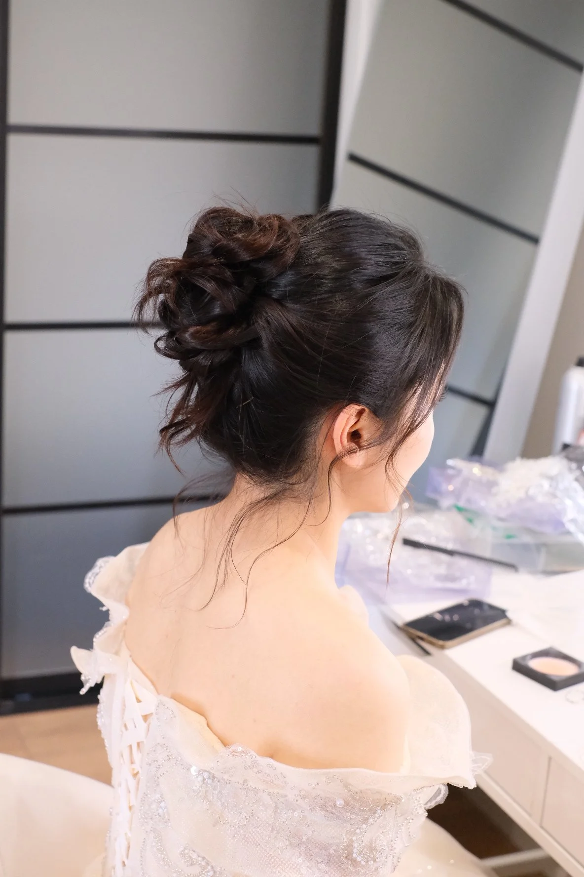 A woman with dark brown hair styled in an elegant updo, sitting at a dressing table in a white dress, with various makeup and beauty items on the table in the background.