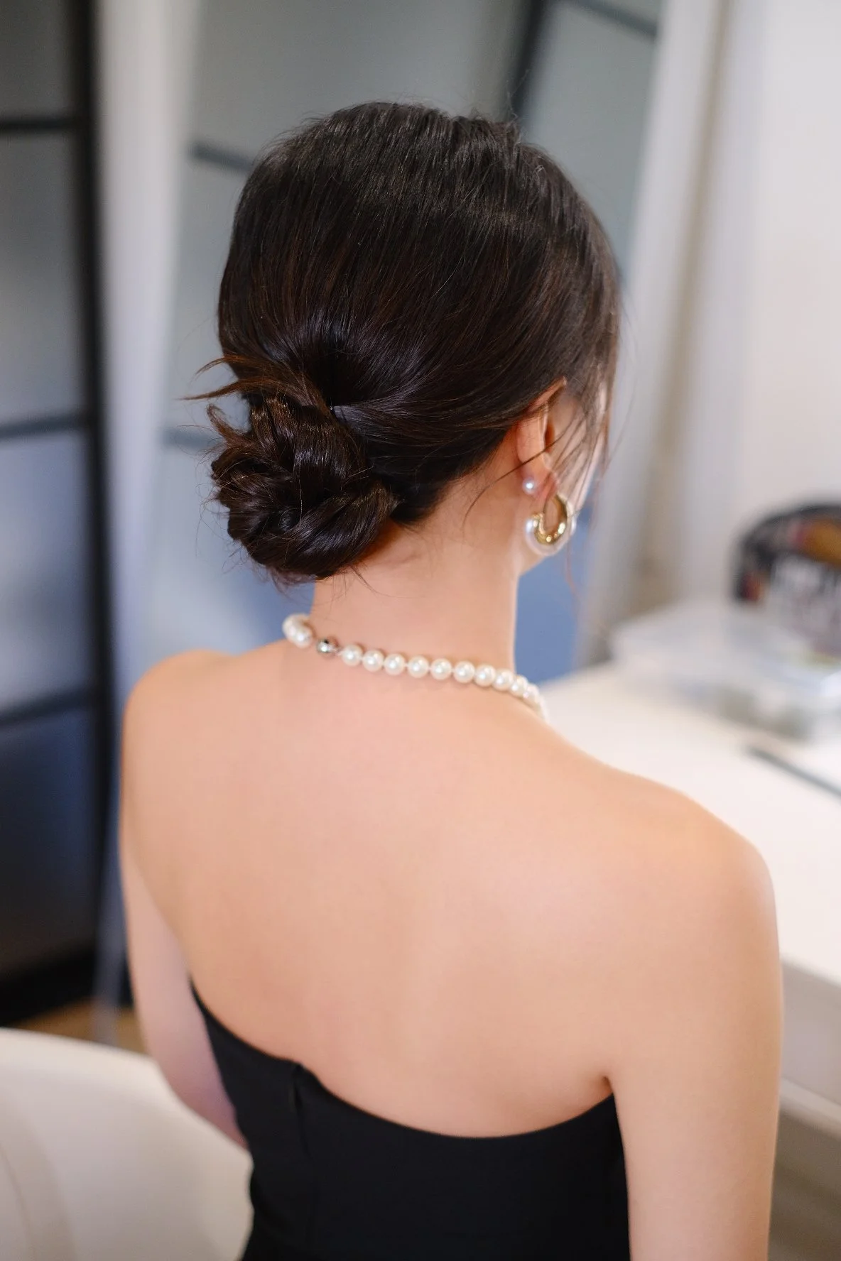 Back of a woman with dark hair styled in an elegant bun, wearing pearl earrings and a pearl necklace, dressed in black, with her bare shoulder visible.