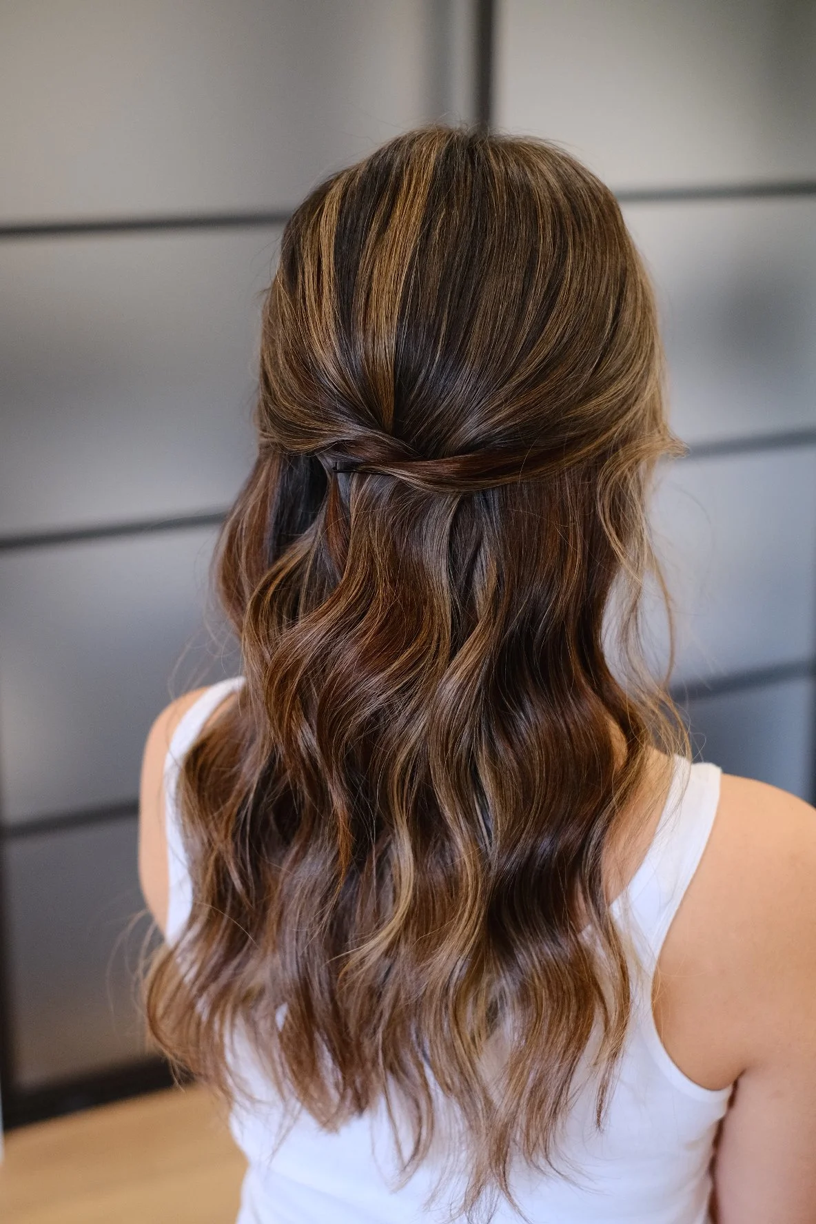 Woman with long wavy hair styled with a twisted half-updo, wearing a white tank top, standing against a modern gray wall.