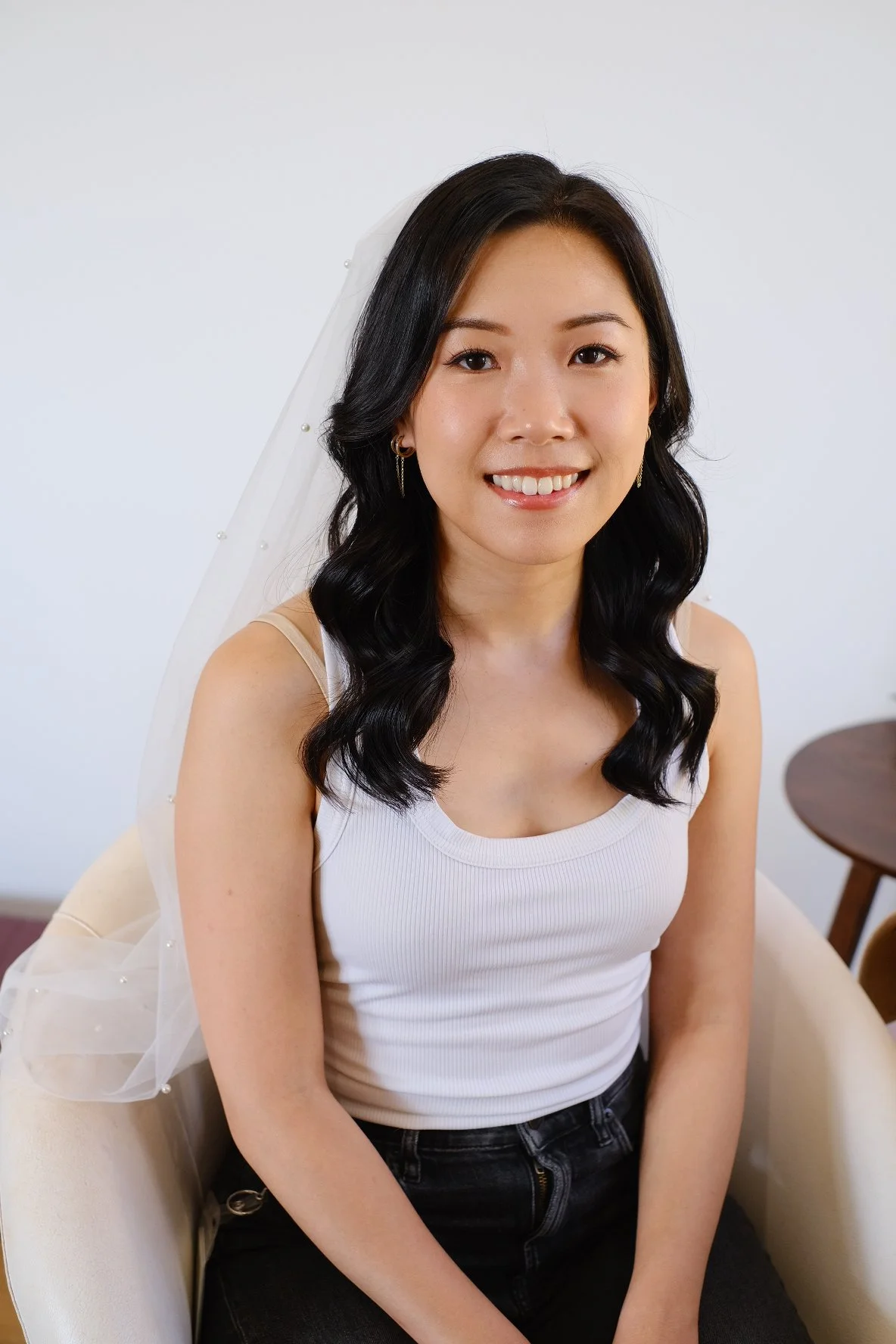 A young woman with dark wavy hair wearing a white sleeveless top, smiling and sitting on a beige chair with a veil in the background.