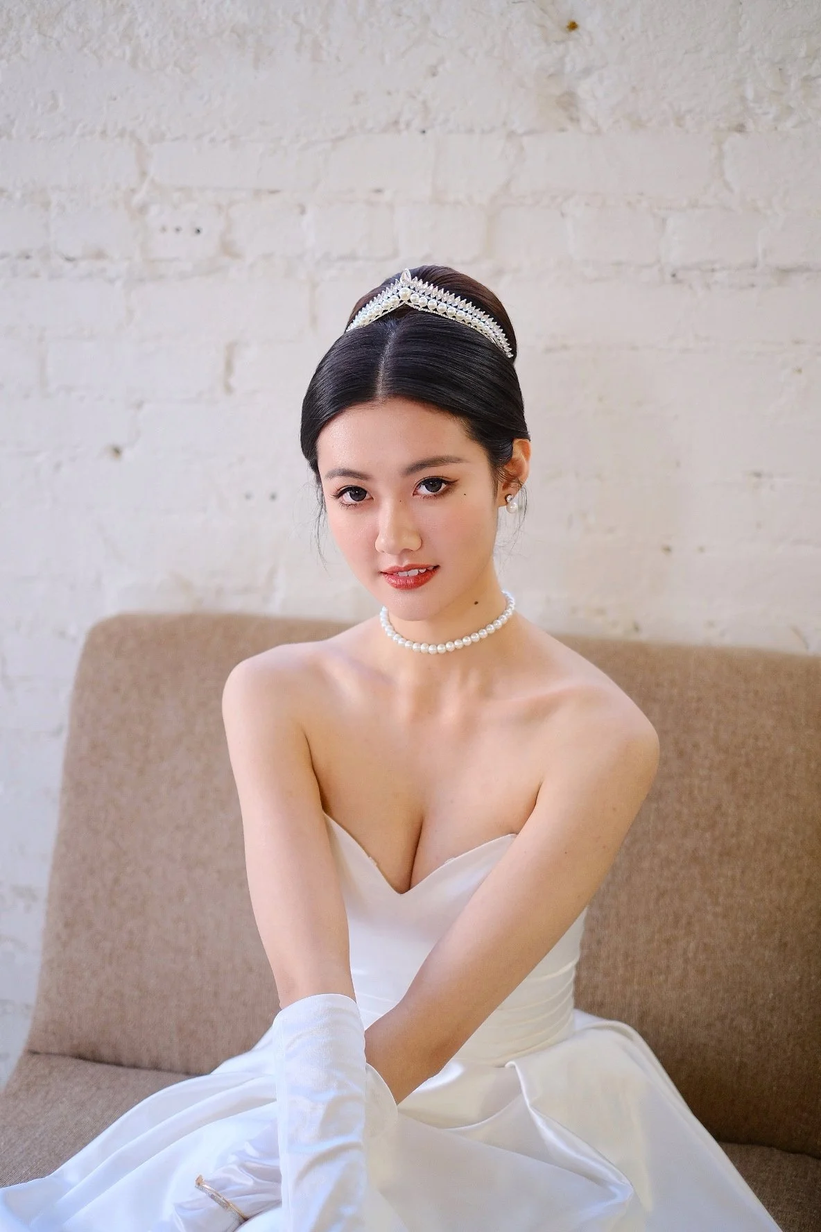 A young woman in a white strapless dress with pearl jewelry, sitting on a beige sofa against a white brick wall, looking at the camera.