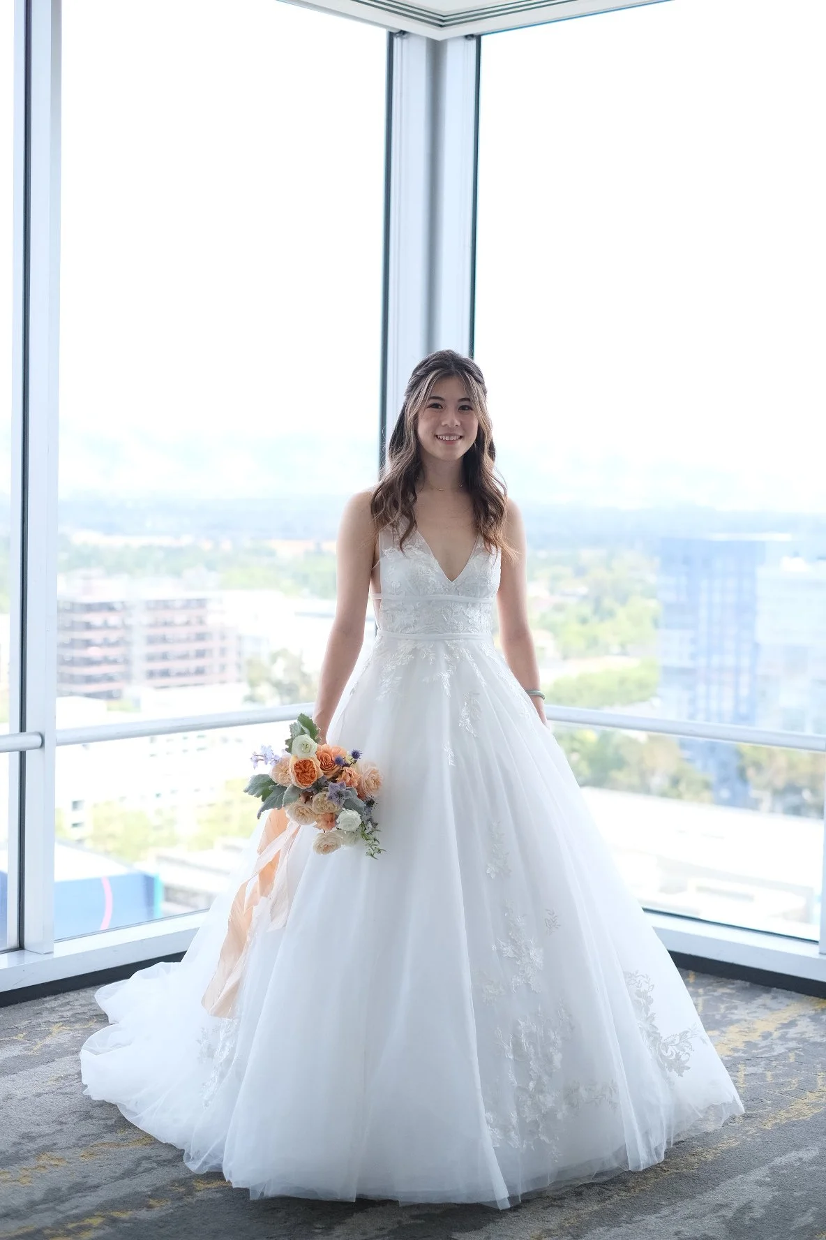 A woman in a white wedding dress holding a bouquet of flowers, standing inside a high-rise building with large windows and cityscape view in the background.