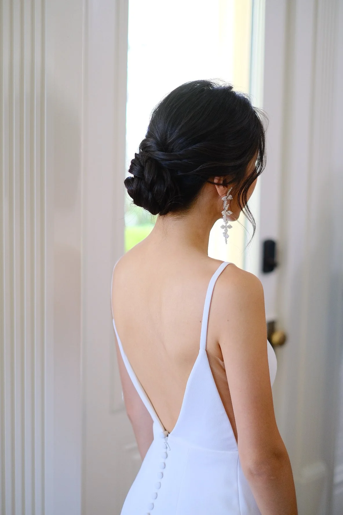 Woman in a white dress with a low back and buttons, her dark hair styled in an elegant bun, standing near a door.
