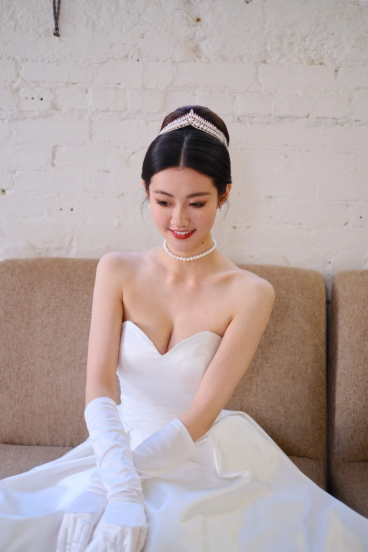 A young woman dressed in a white strapless wedding gown, wearing a pearl necklace, pearl earrings, a tiara, and long white gloves, sitting on a beige couch in front of a white brick wall.