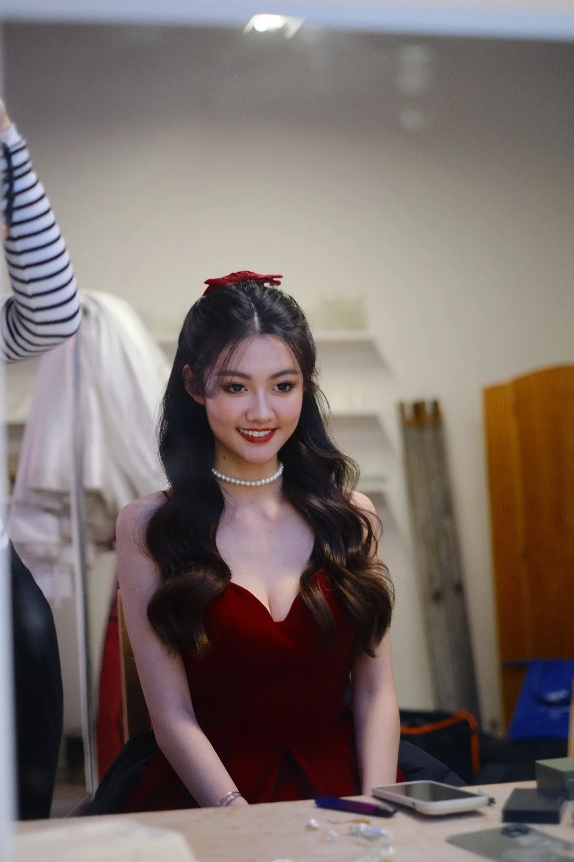 A young woman with long dark wavy hair, wearing a red dress and a pearl necklace, smiling indoors.