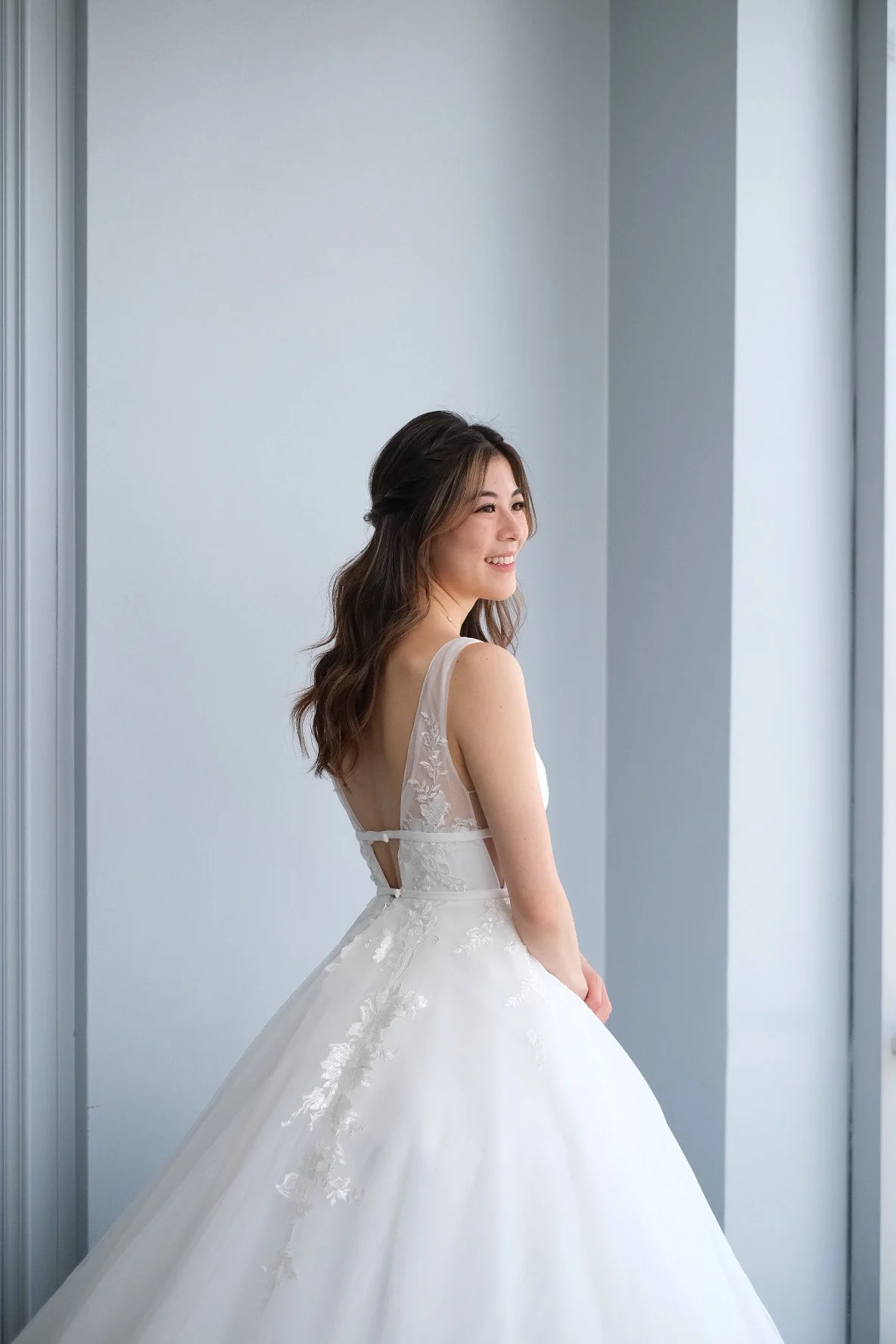 A woman in a white wedding dress with lace details standing near a window and smiling.