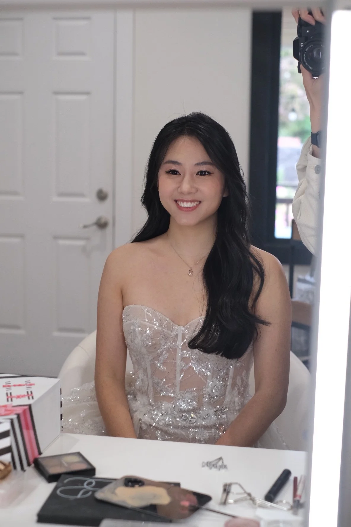A young woman with long black hair smiling while sitting in front of a mirror, wearing a strapless, glamorous dress with sequins and tulle. A person holding a camera is visible in the mirror reflection.