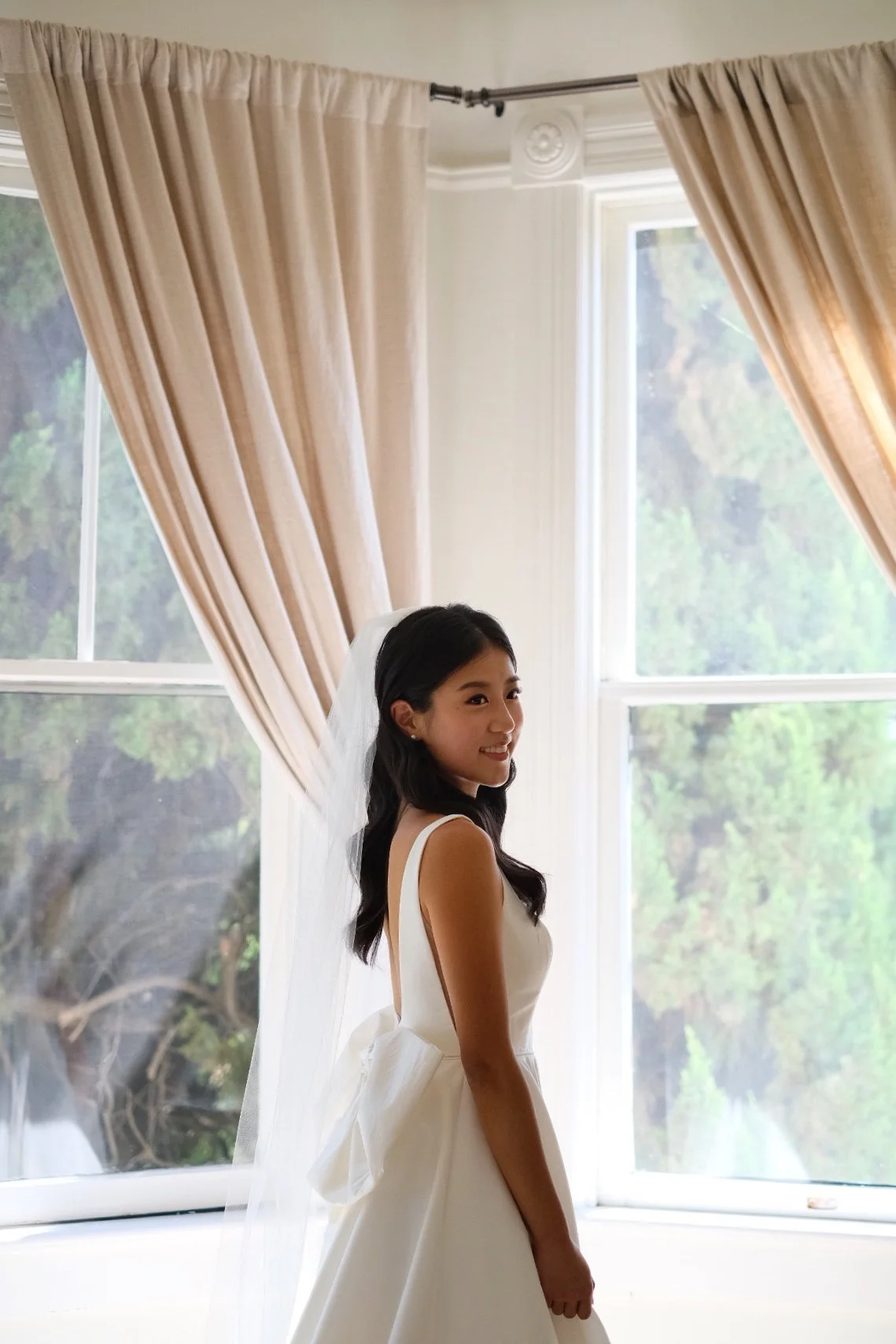 A woman is dressed in a wedding gown with a veil, standing by a large window with beige curtains. Trees are visible outside the window.