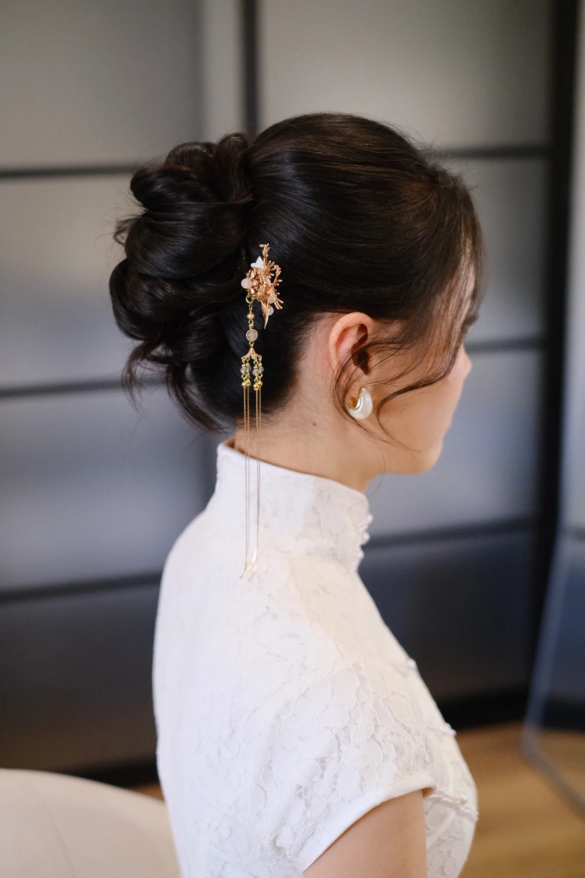A woman with dark hair styled in an elegant updo, adorned with a large gold and pearl hair accessory, wearing pearl earrings and a high-collared white lace dress, standing indoors against a gray background.