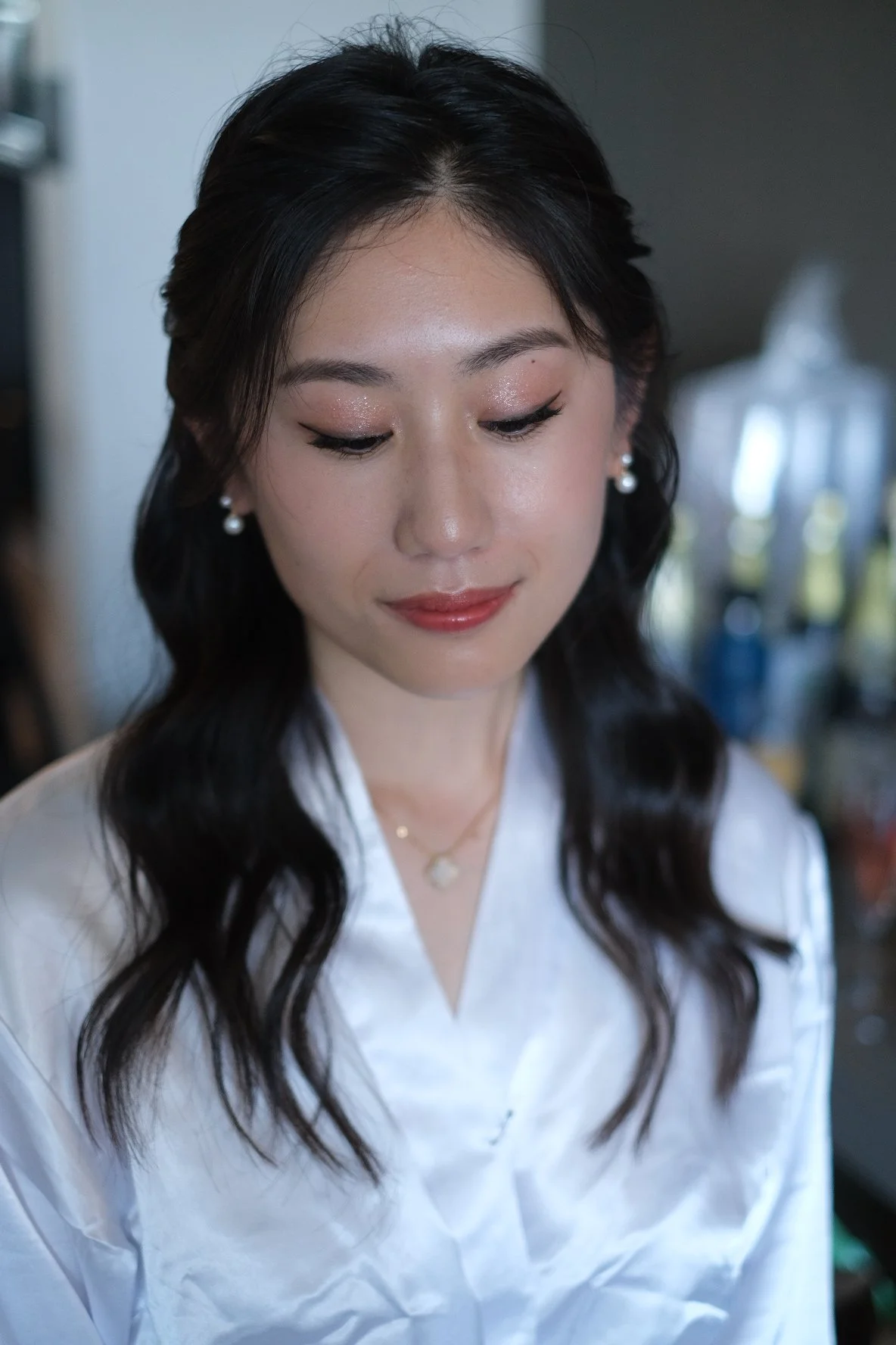 A young woman with long dark hair styled in loose waves, wearing pearl earrings and a delicate necklace, dressed in a white blouse. She has makeup with glittery eyeshadow and red lipstick, and her eyes are looking downward with a soft smile.